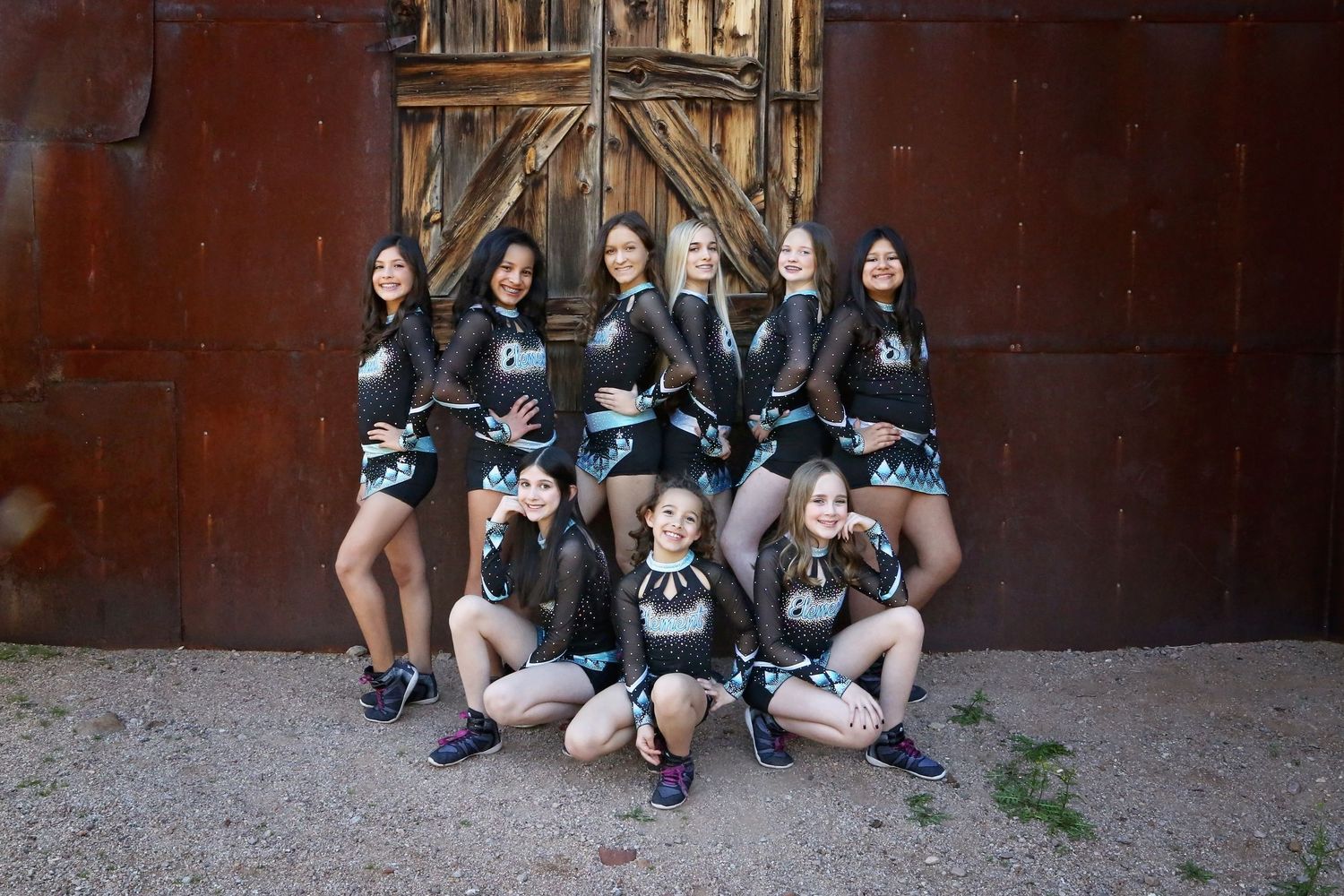 Cheer team in black and teal uniforms poses in rows in front of a rustic wooden barn door and rusty metal wall.