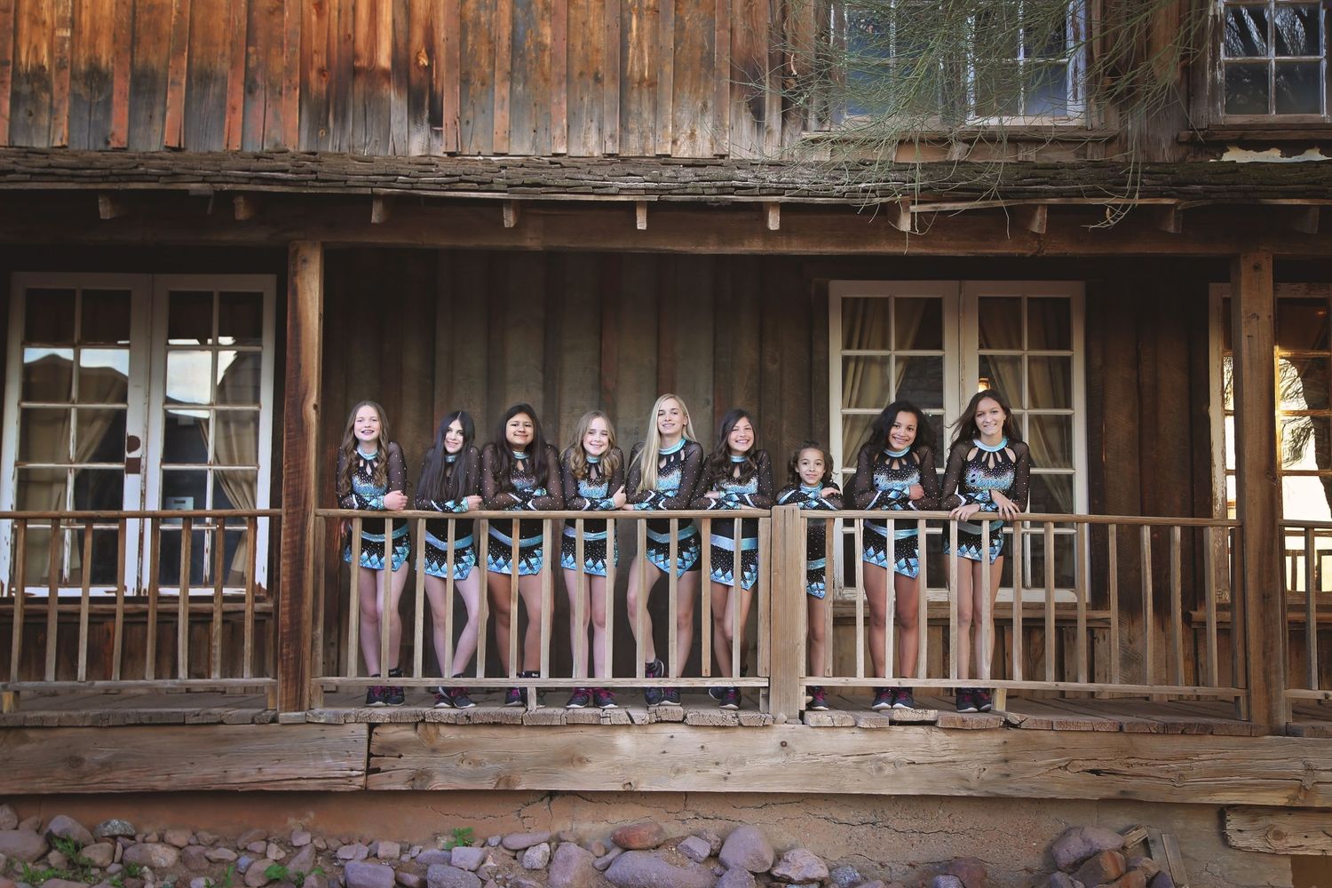 Cheer team in matching black and teal uniforms poses together on the porch of a rustic old western wooden building.