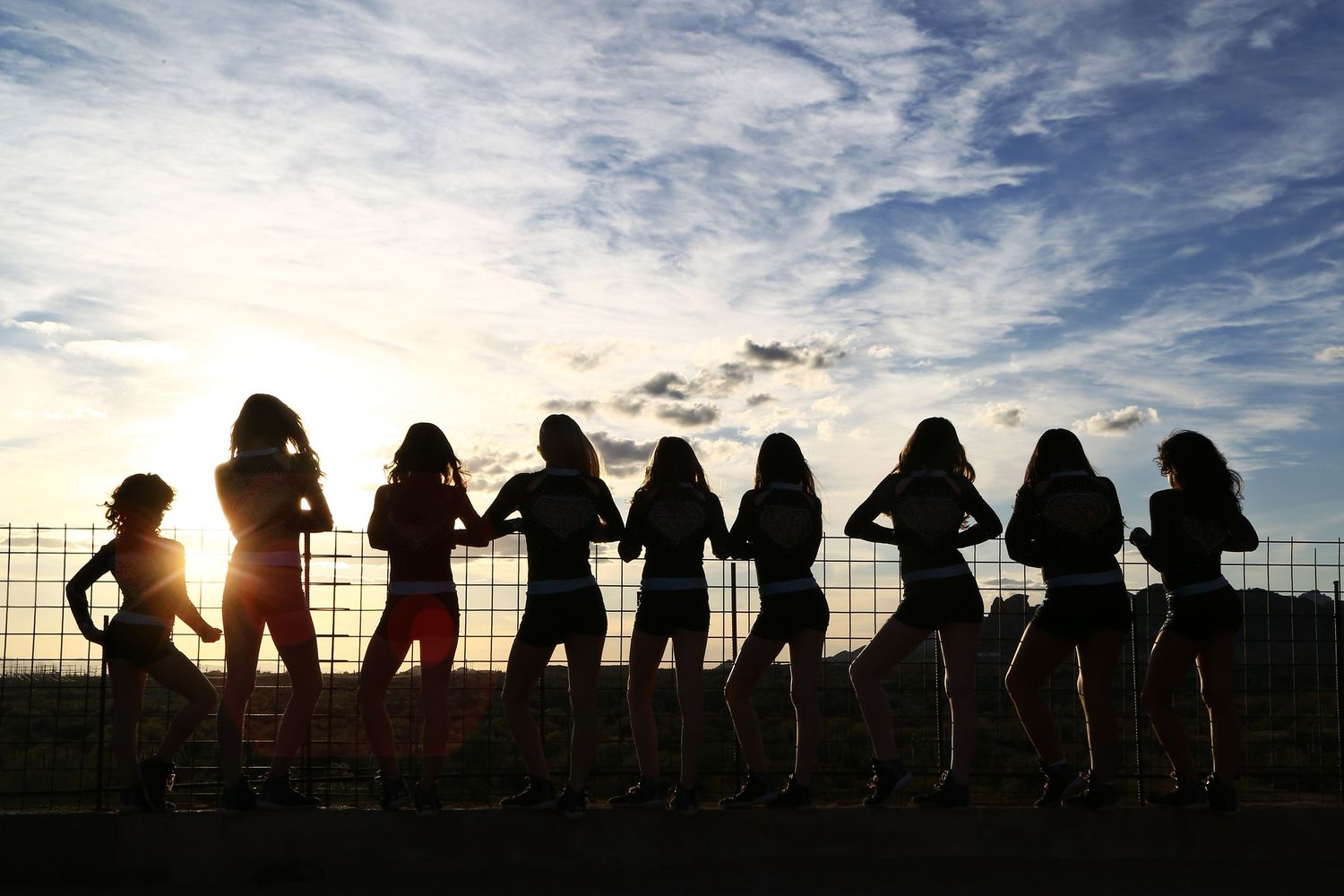Silhouettes of cheerleaders lined up along a fence at sunset with a bright sun and wispy clouds in the sky.