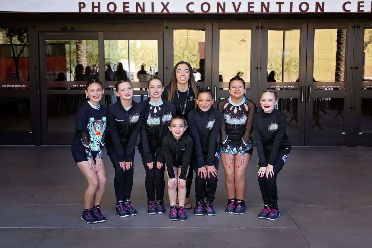 Youth cheerleaders in black uniforms pose together in front of the Phoenix Convention Center entrance.