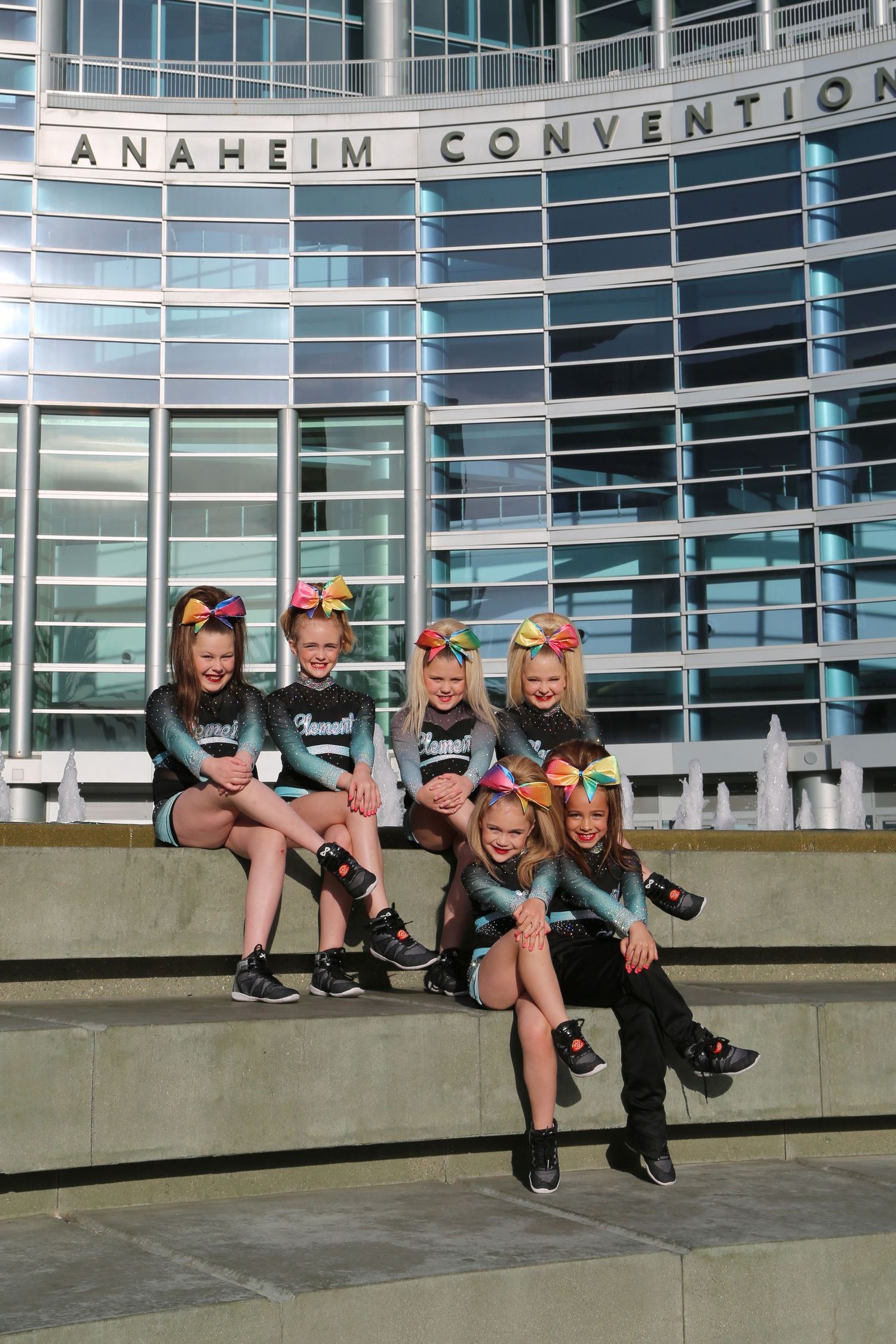 Young cheerleaders with colorful bows sit on steps outside the Anaheim Convention Center.