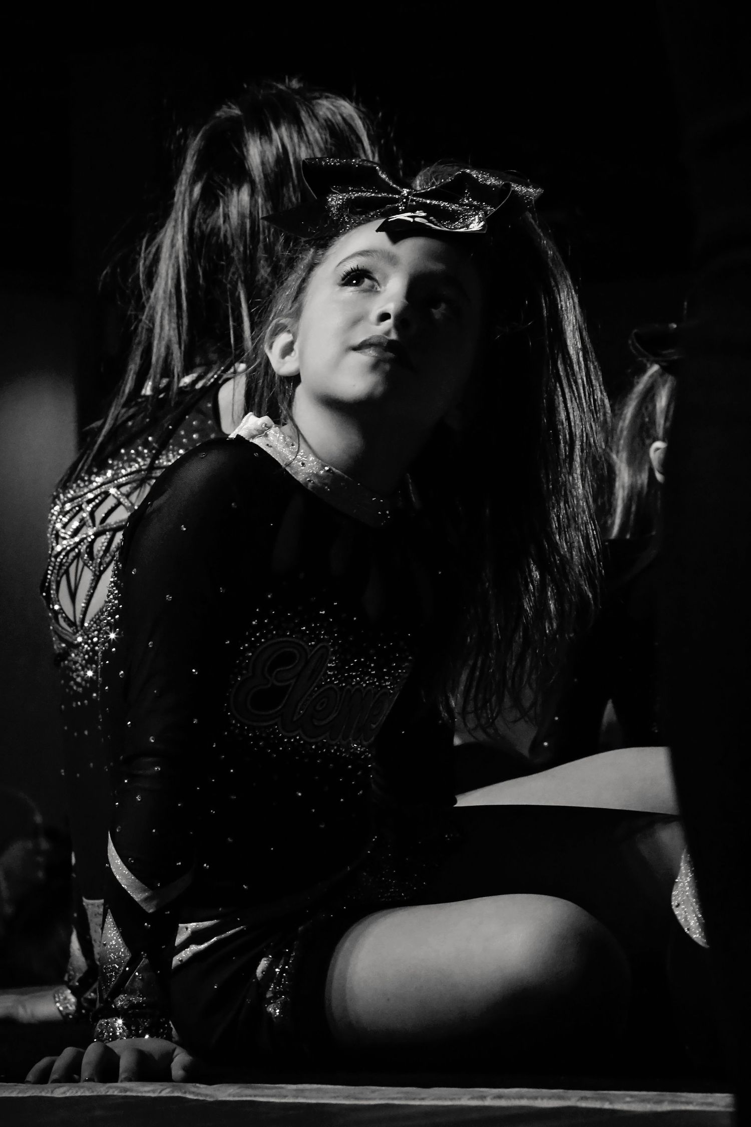Black and white photo of a cheerleader in a sparkly uniform and bow sitting on the floor and gazing upward.