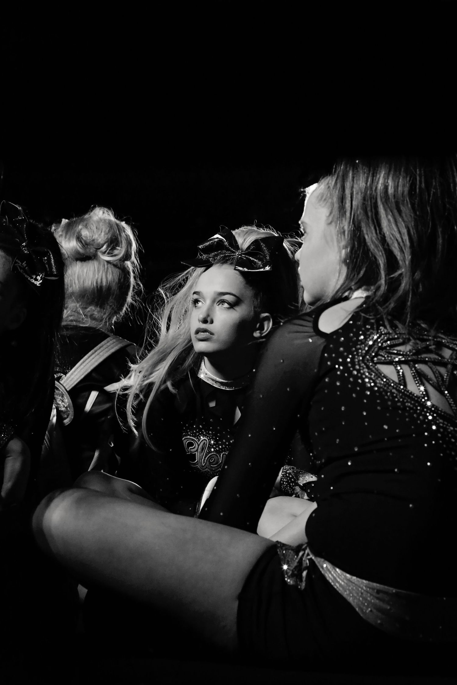 Black and white portrait of a young cheerleader with a large bow glancing sideways, surrounded by teammates in dark sparkling uniforms.