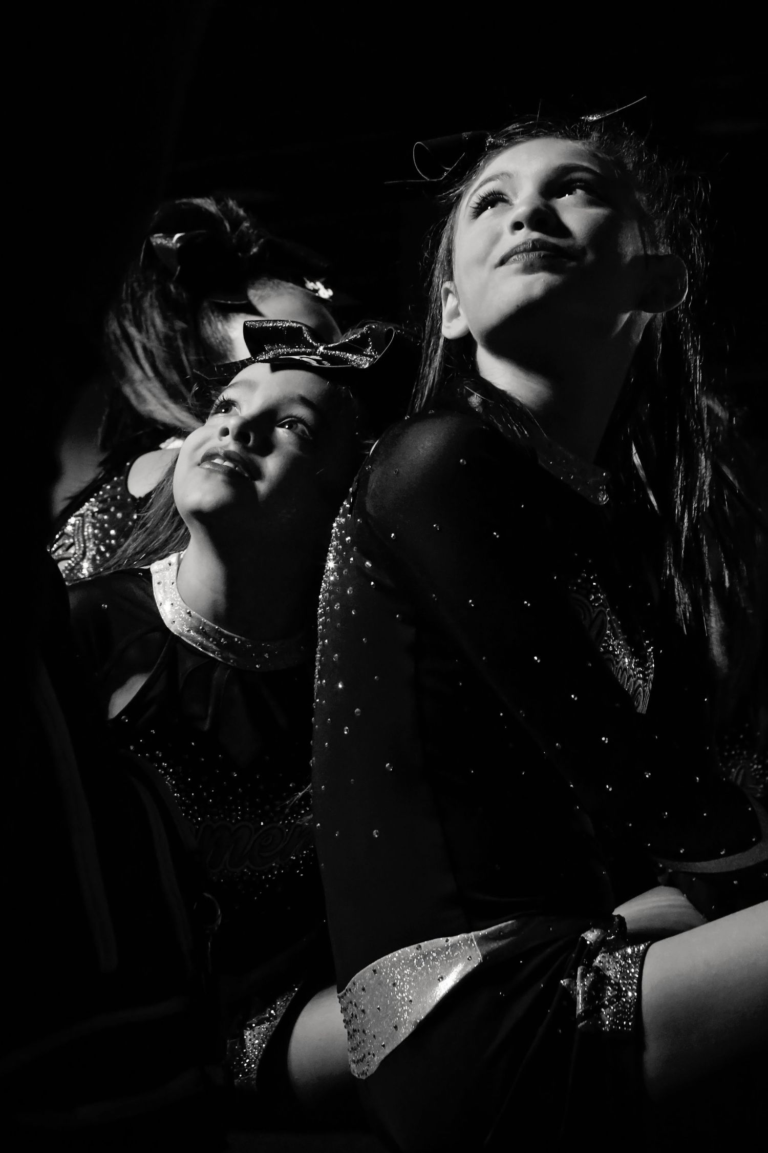 Two cheerleaders in sparkly uniforms look upward with hopeful smiles in dramatic black and white lighting.