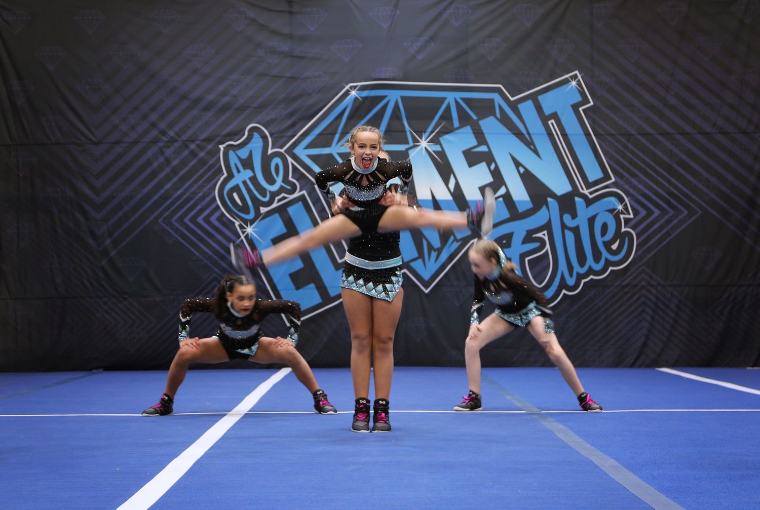 Cheerleaders perform on a blue competition mat as a flyer does a toe touch jump in front of an Elite banner.