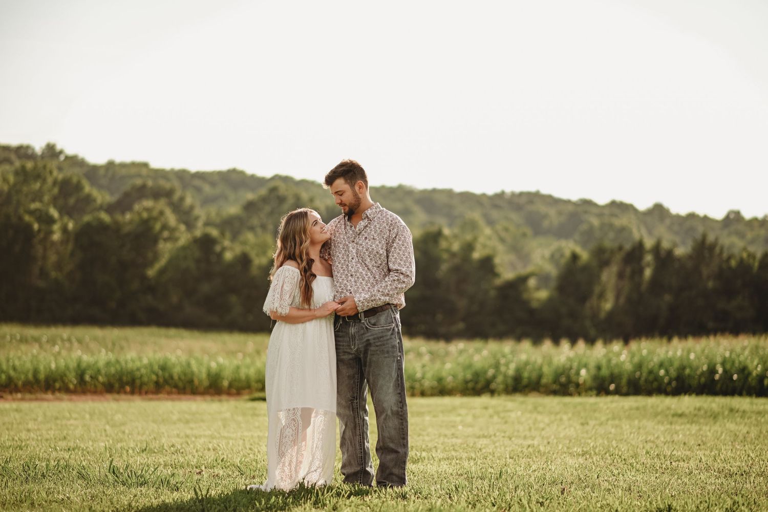 Couple standing together in a lush green farm field with corn rows and trees in the background.