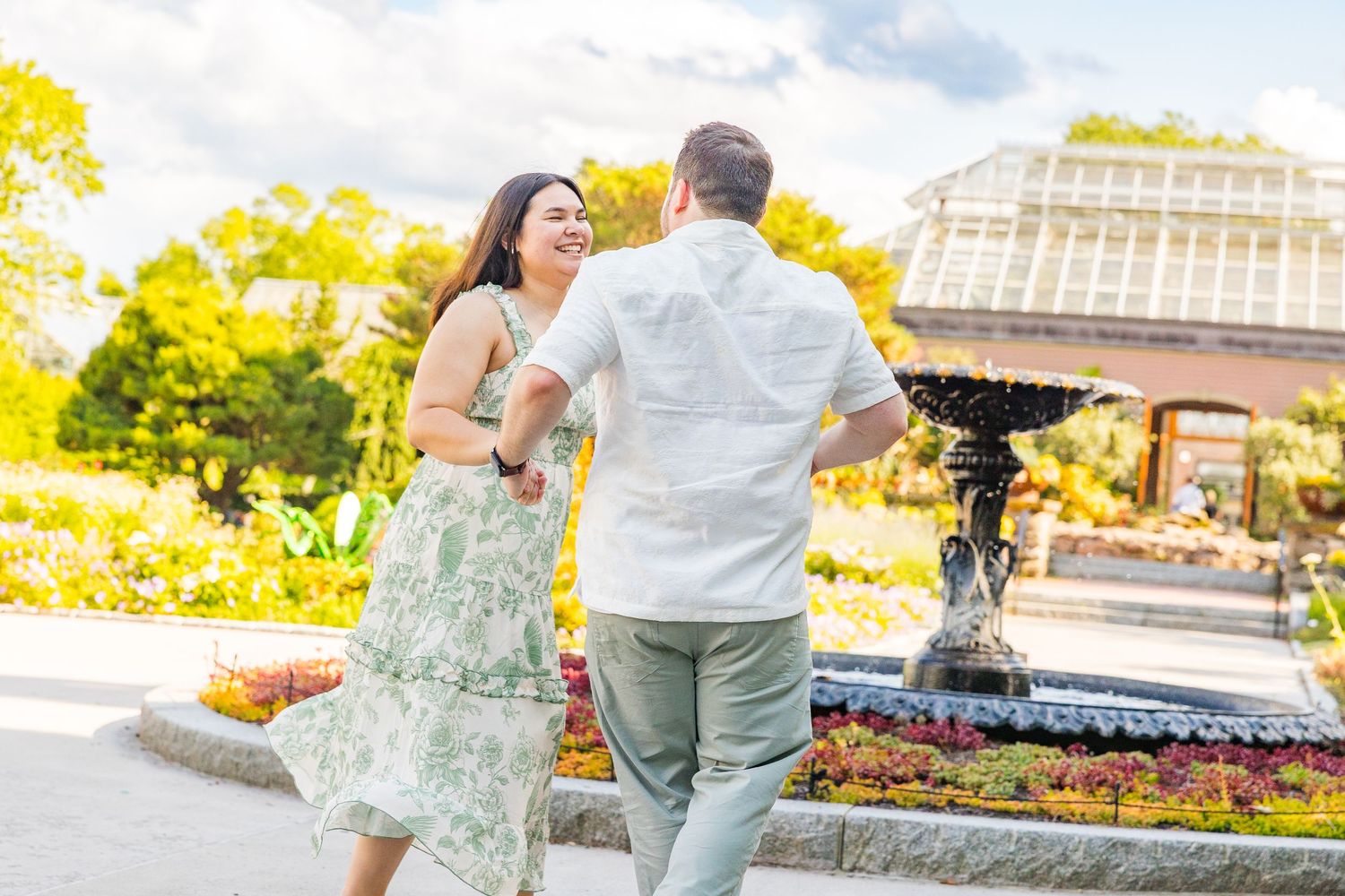 Couple dancing and laughing together in a beautiful garden with a fountain and greenhouse backdrop.