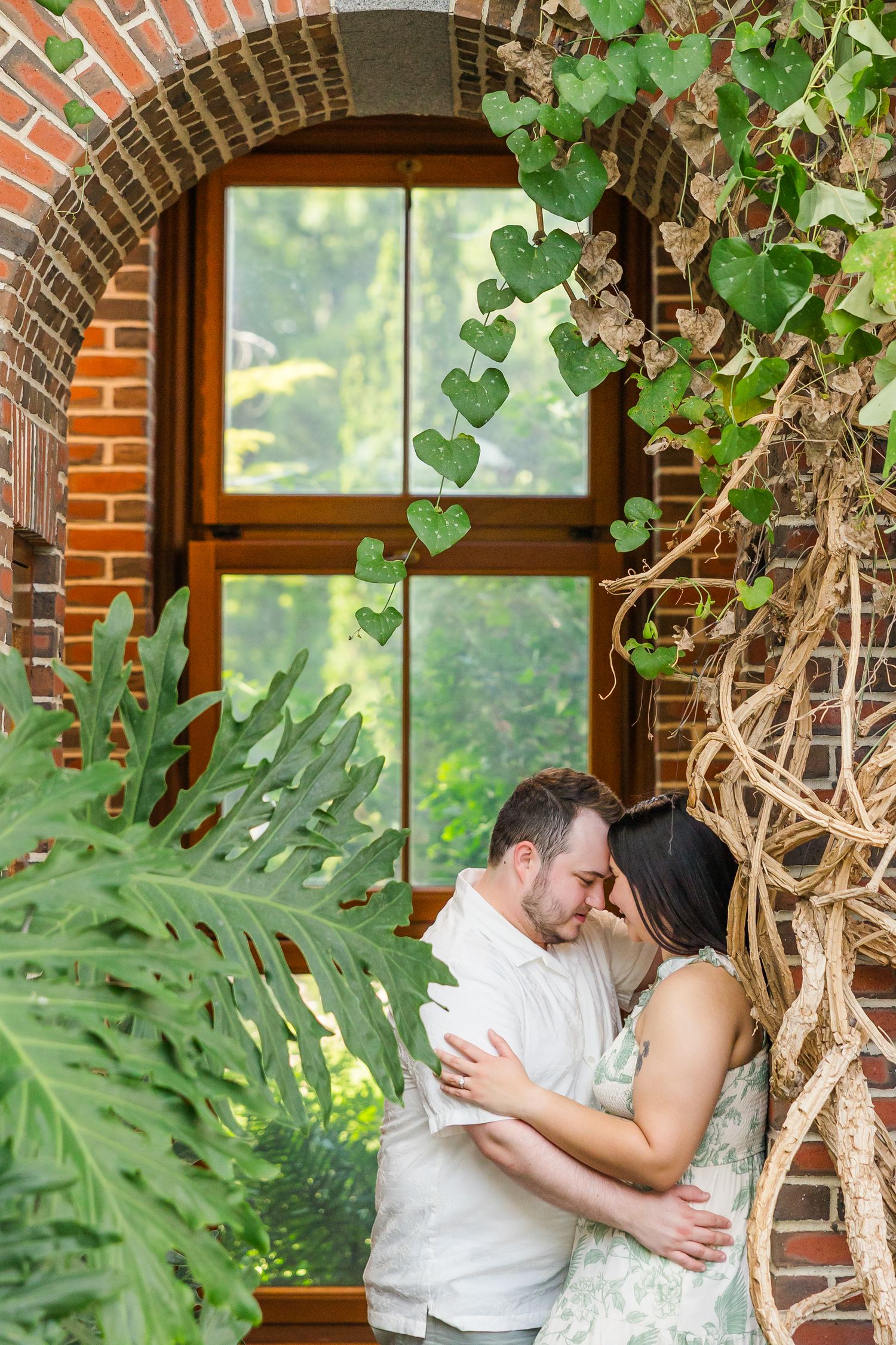 Couple embracing tenderly under a brick archway surrounded by lush green ivy and tropical plants near a wooden window.