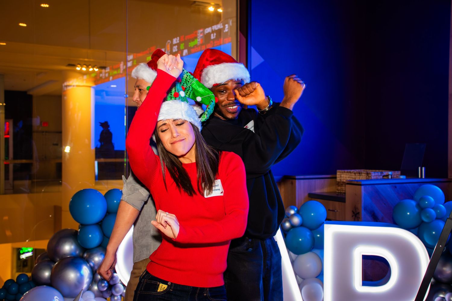 Two people wearing Santa hats dance joyfully at a festive holiday party with blue balloons and decorations.