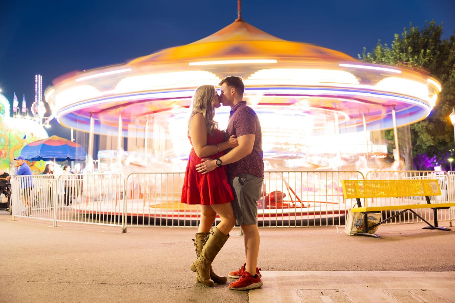 Couple embracing romantically in front of a glowing carousel at a colorful amusement park at night.