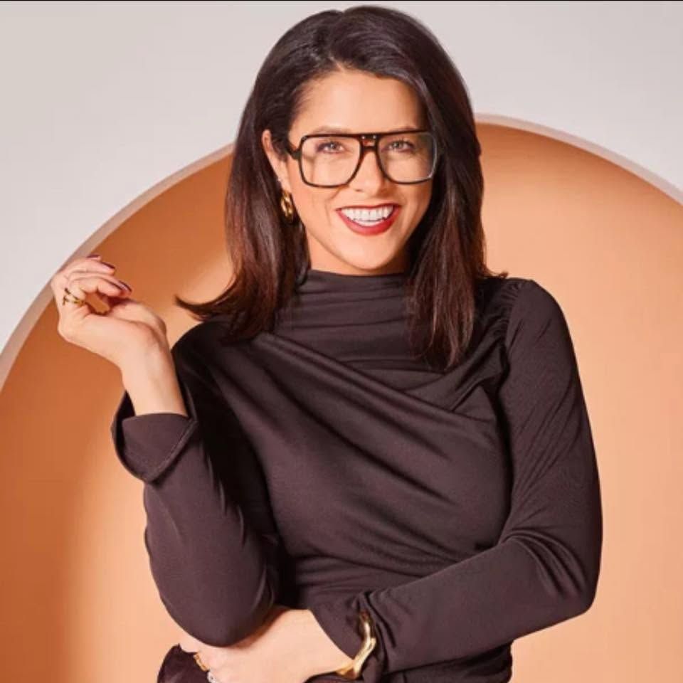 Smiling woman with dark hair and bold square glasses wearing a dark turtleneck top, posing confidently in front of a circular backdrop.