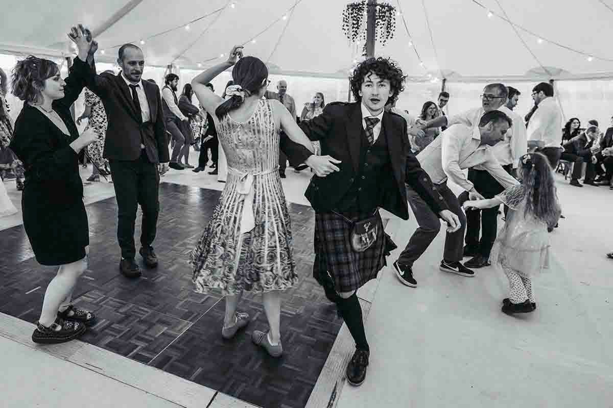 Black and white photo of guests dancing energetically at a wedding ceilidh with a young man in a kilt at a private marque wedding in Penrith