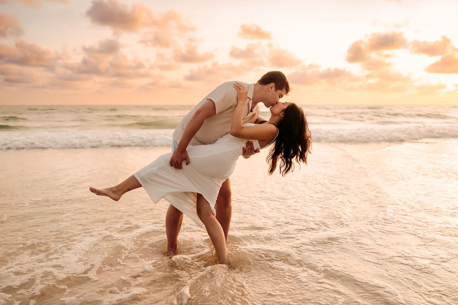 Romantic couple sharing a passionate dip kiss on Anna Maria Island at golden sunset with waves gently washing around their feet.
