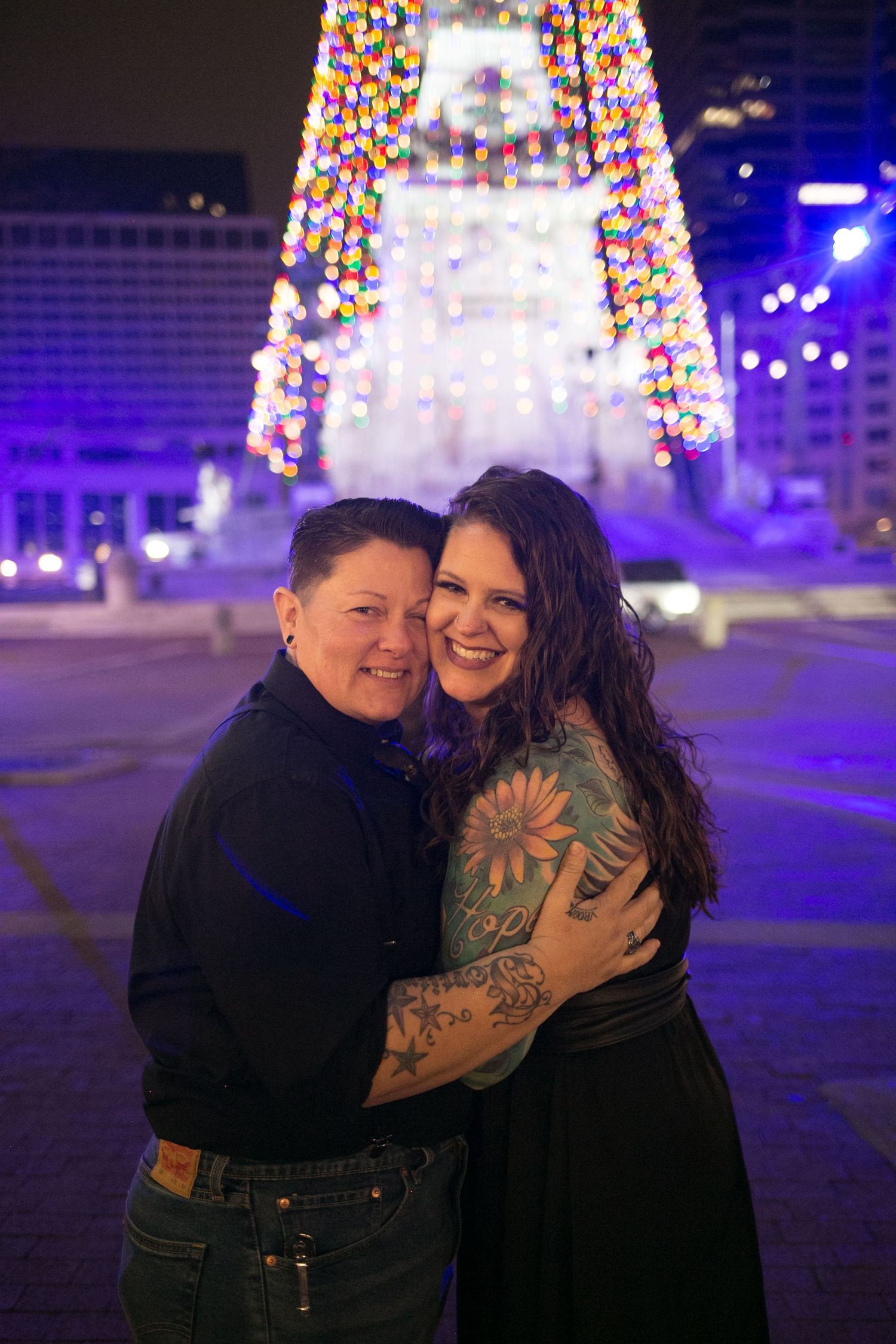 Two women embracing and smiling in front of a lit holiday monument at night, one with colorful tattoos on her arm.
