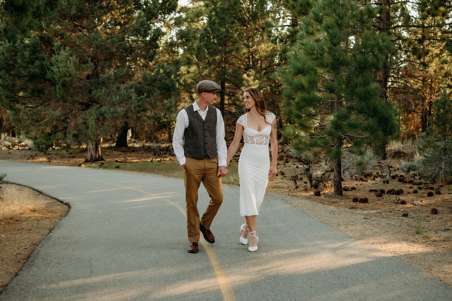 Couple holding hands smiling at each other against a red brick wall, wearing elegant attire.