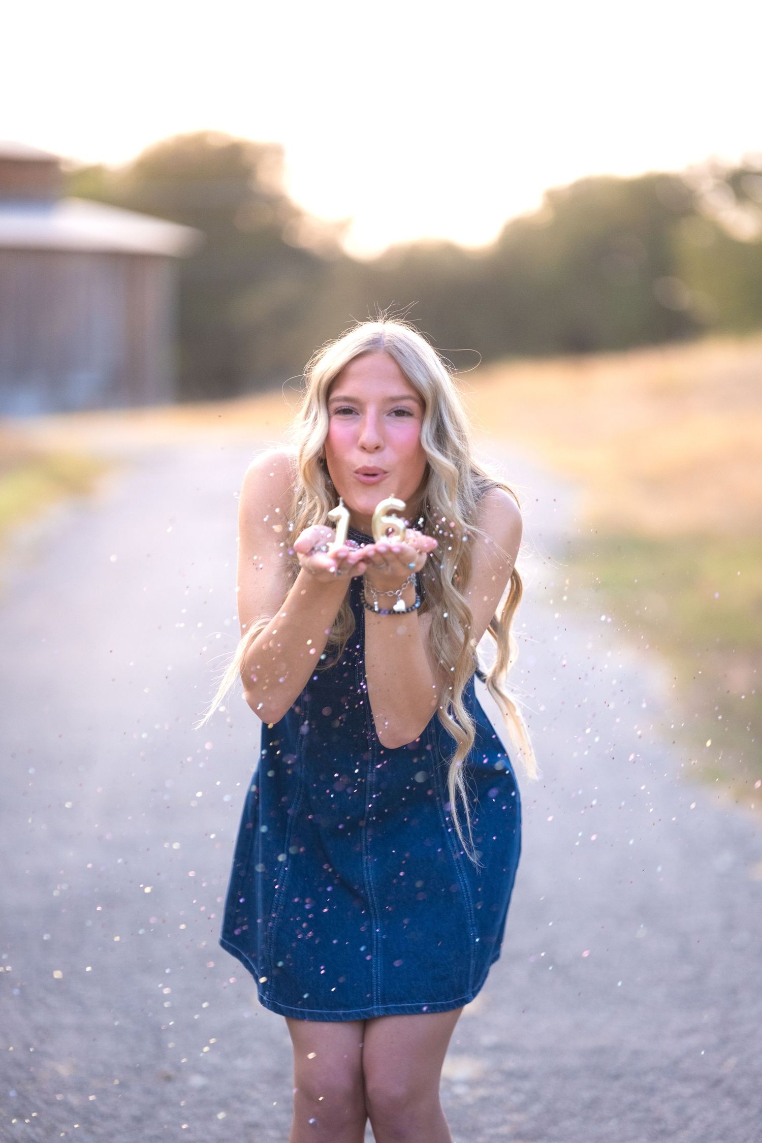 Blonde woman blowing glitter in golden sunset light on rural path for sweet 16 birthday celebration at Salado Creek Pace Park Salado Texas