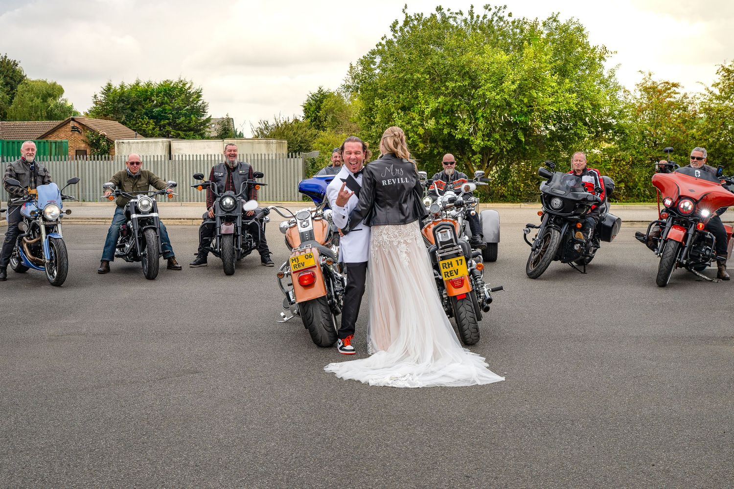 Bride and groom in leather jackets pose with Harley-Davidson motorcycles and bikers in a parking lot on their wedding day.