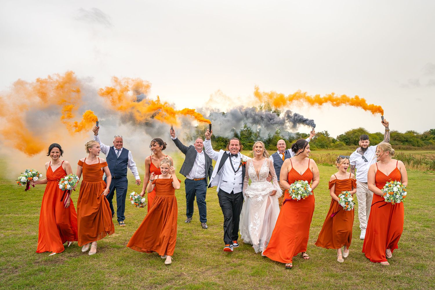 Wedding party walks joyfully outdoors with orange smoke bombs, bridesmaids in rust dresses holding white bouquets.Yorkshire, Sheffield, UK