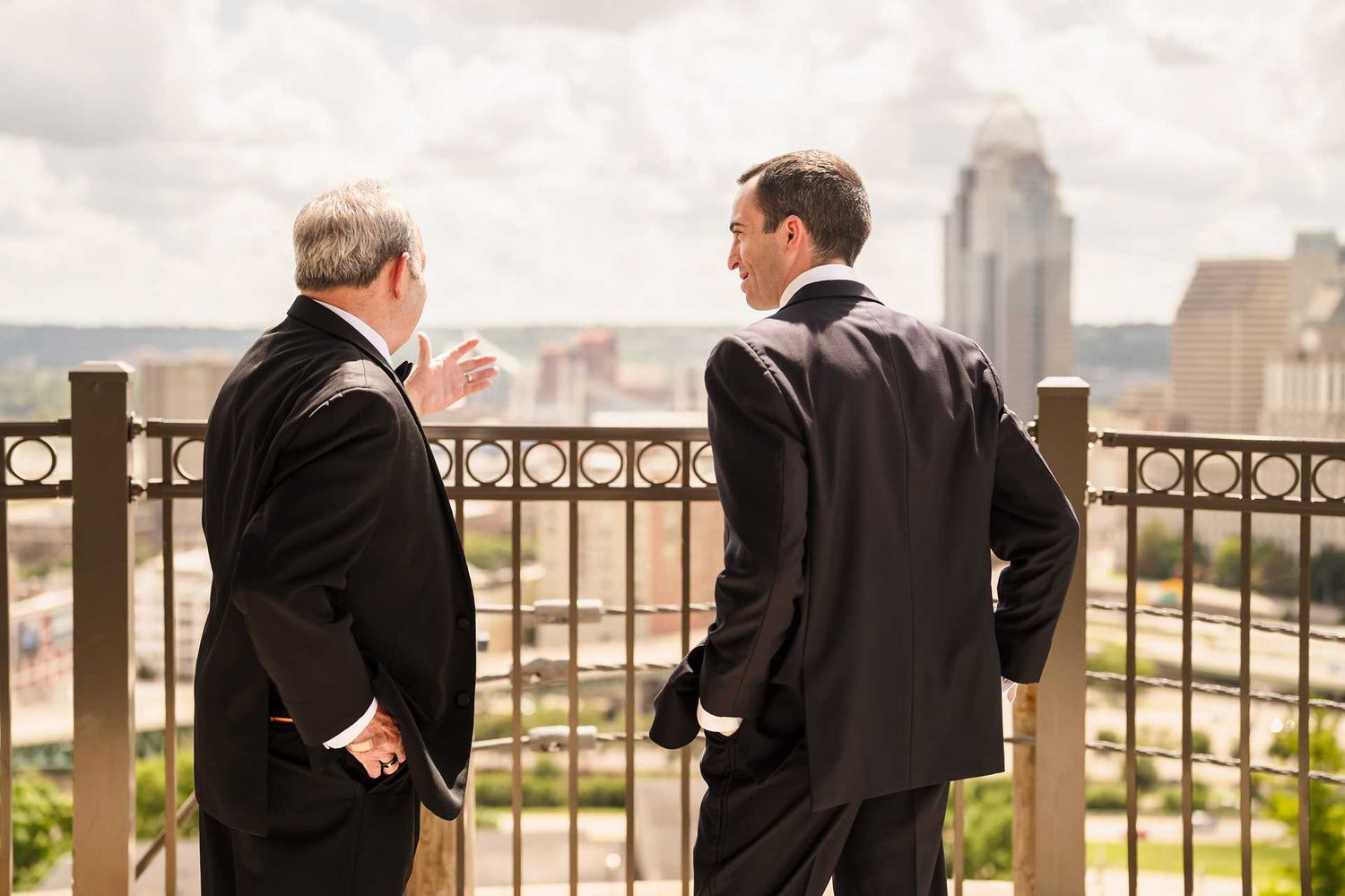 Two men in dark suits converse on an outdoor balcony overlooking a city skyline on a cloudy day.