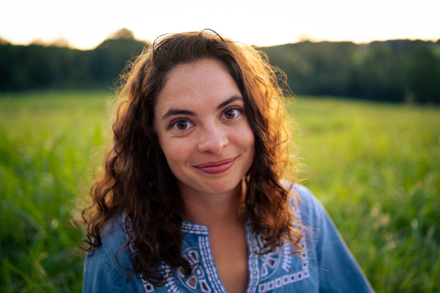 A curly-haired woman smiles softly in a sunlit green field at golden hour.