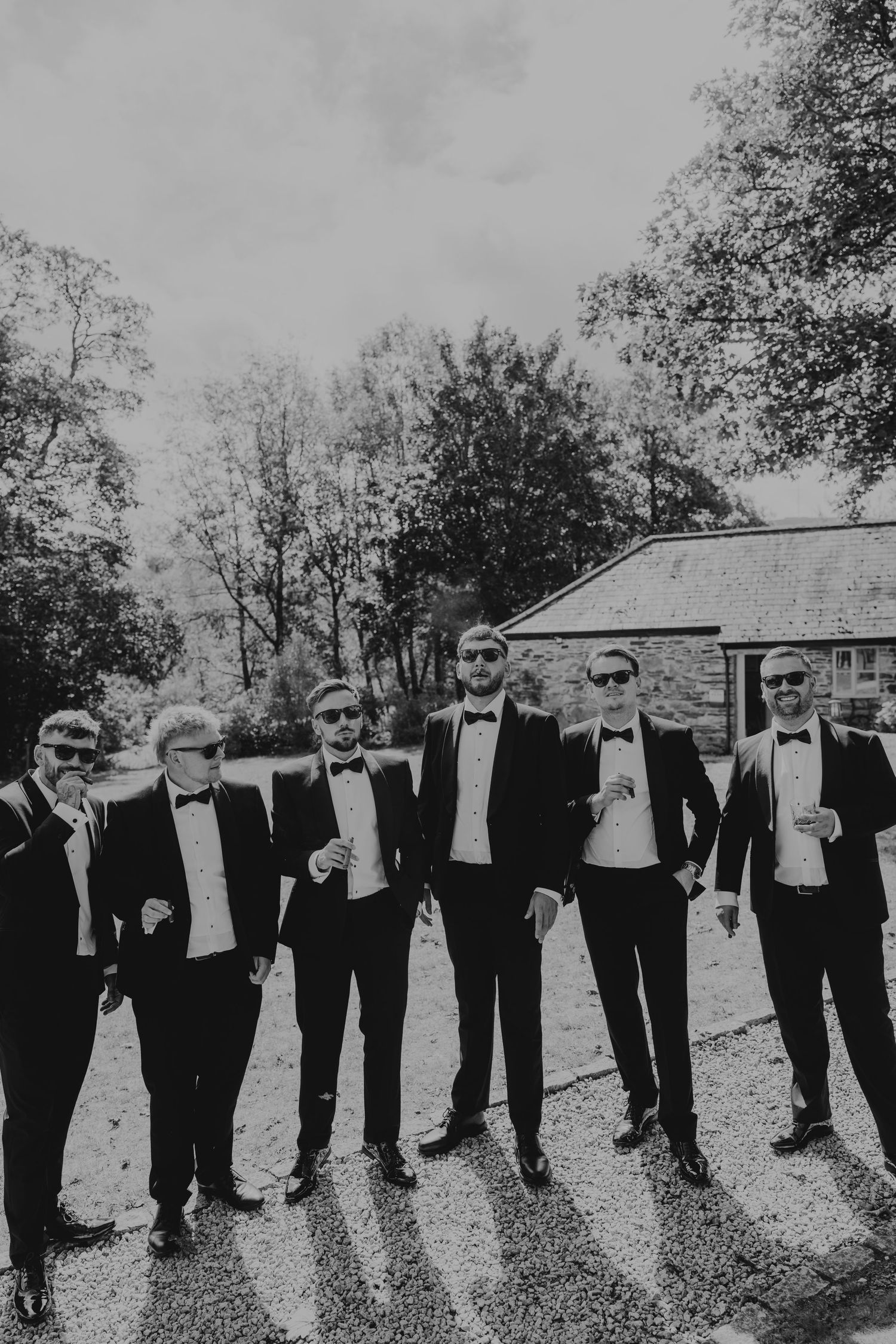 Six groomsmen in black tuxedos and sunglasses pose outdoors on gravel at a wedding venue in a black and white photo.