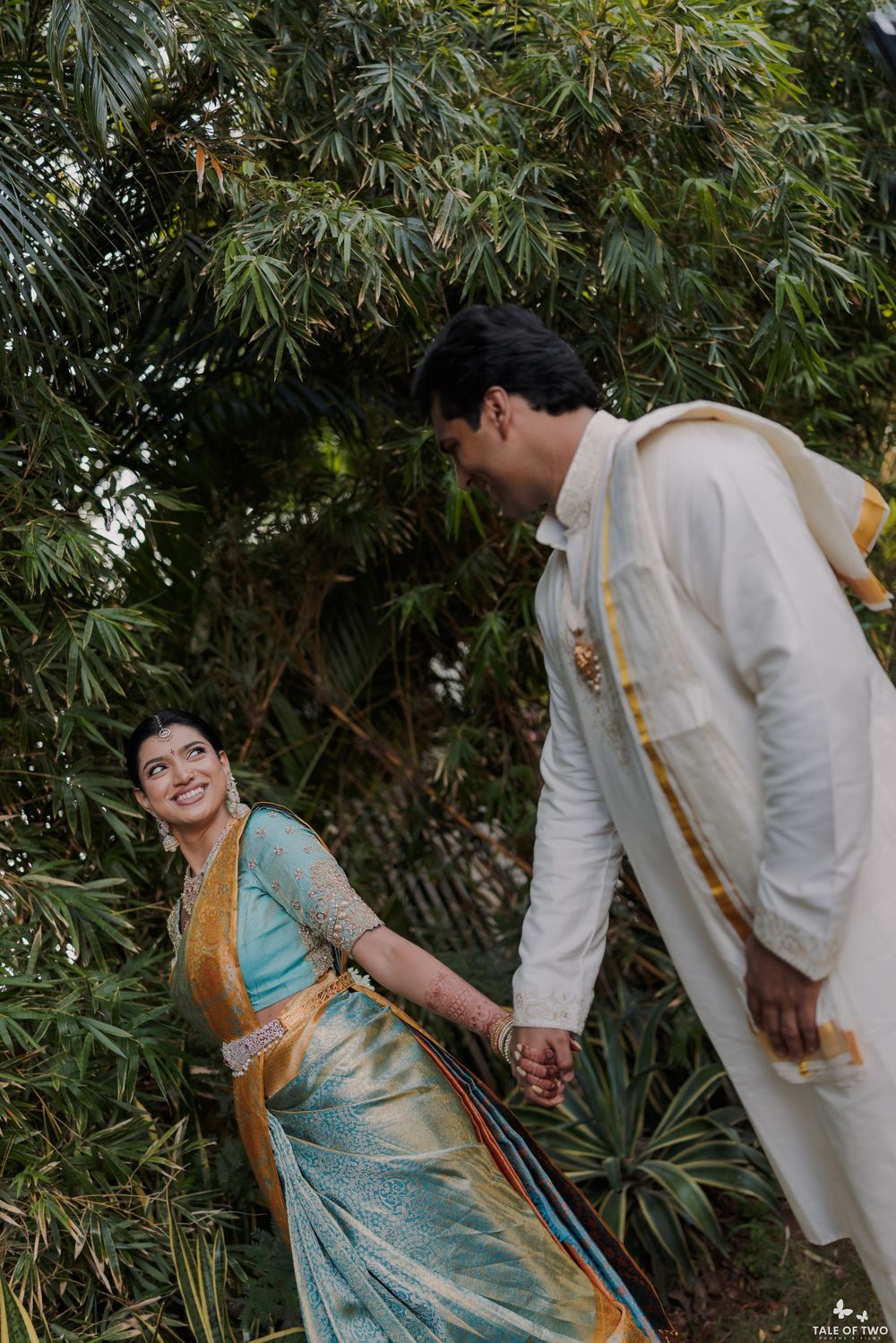 Joyful South Indian bride in teal Kanjivaram saree holds groom's hand amid lush green foliage.