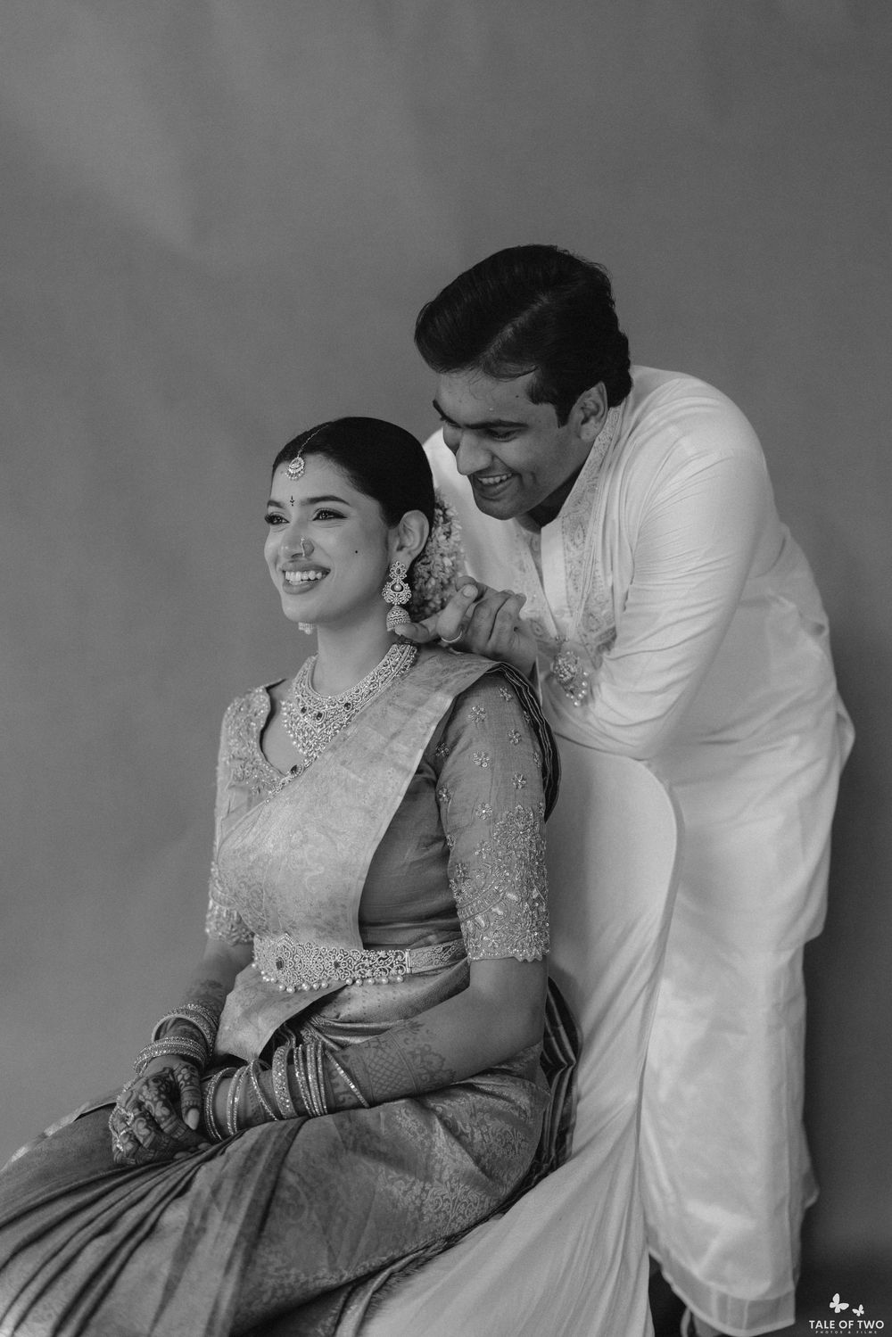 Black and white portrait of smiling Indian bride in saree seated with groom standing behind her.