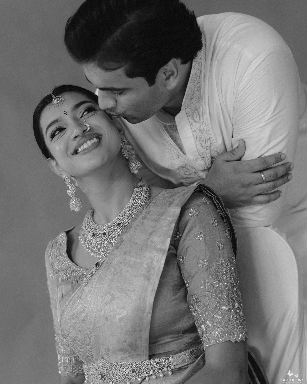 Black and white photo of a couple in traditional Indian attire, man kissing woman on cheek as she smiles joyfully.