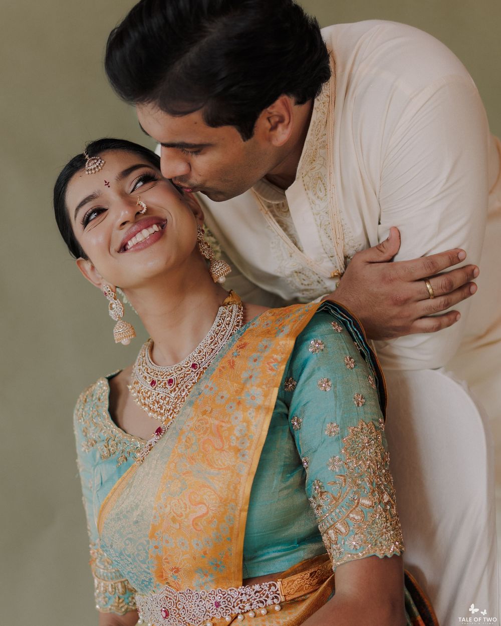 Indian couple in traditional wedding attire, bride in teal saree with gold jewelry, groom kissing her cheek.