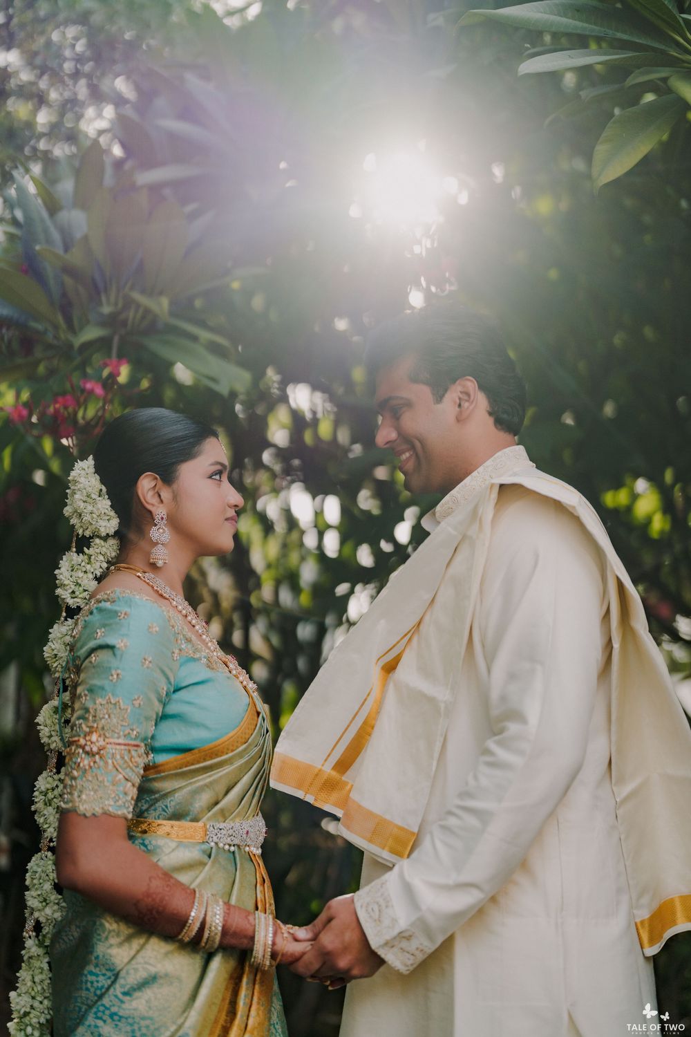 South Indian bride in teal silk saree and groom in white sherwani hold hands in a sunlit garden.