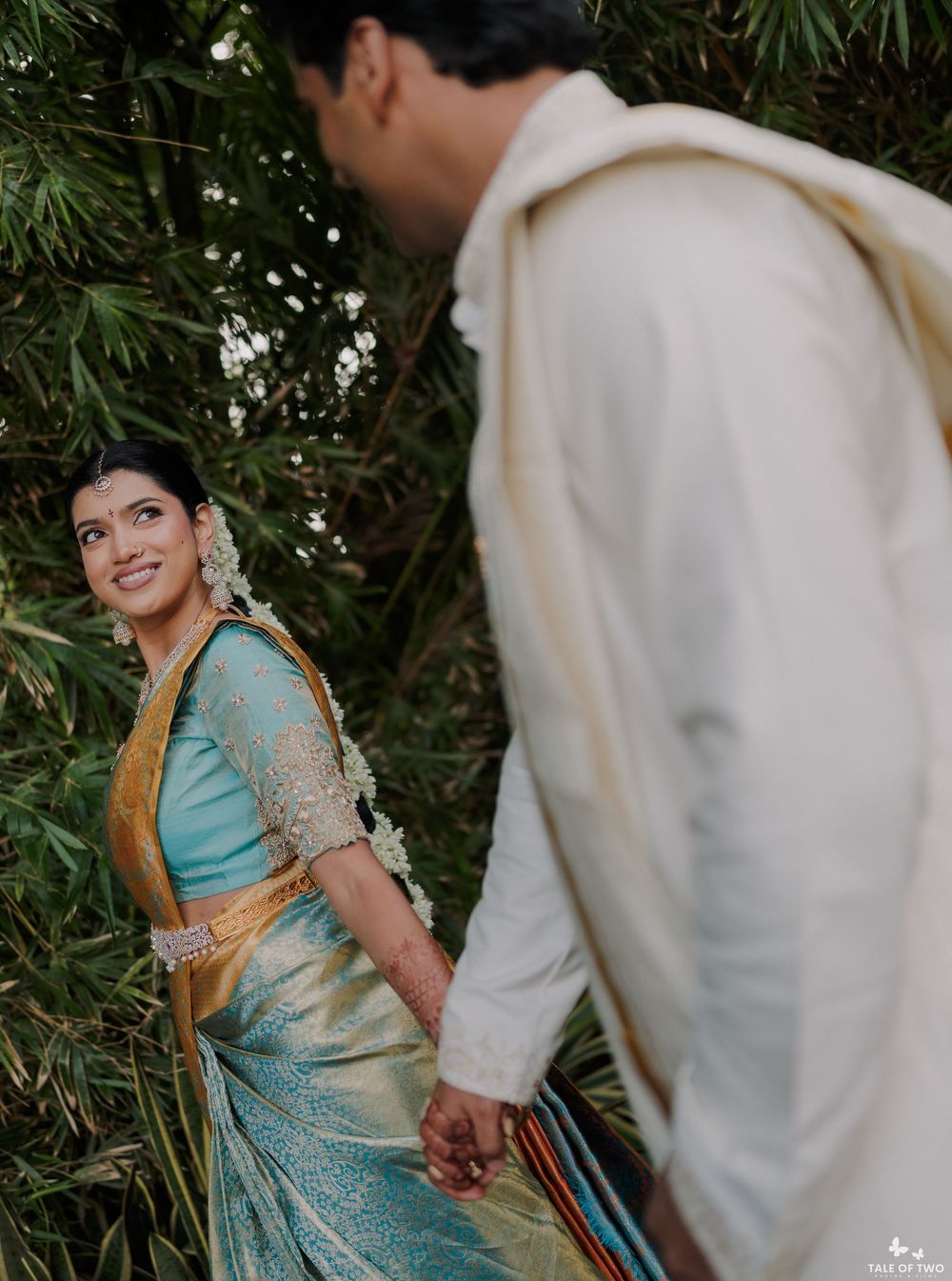 South Indian bride in teal Kanjivaram saree holds groom's hand while looking back smiling outdoors.