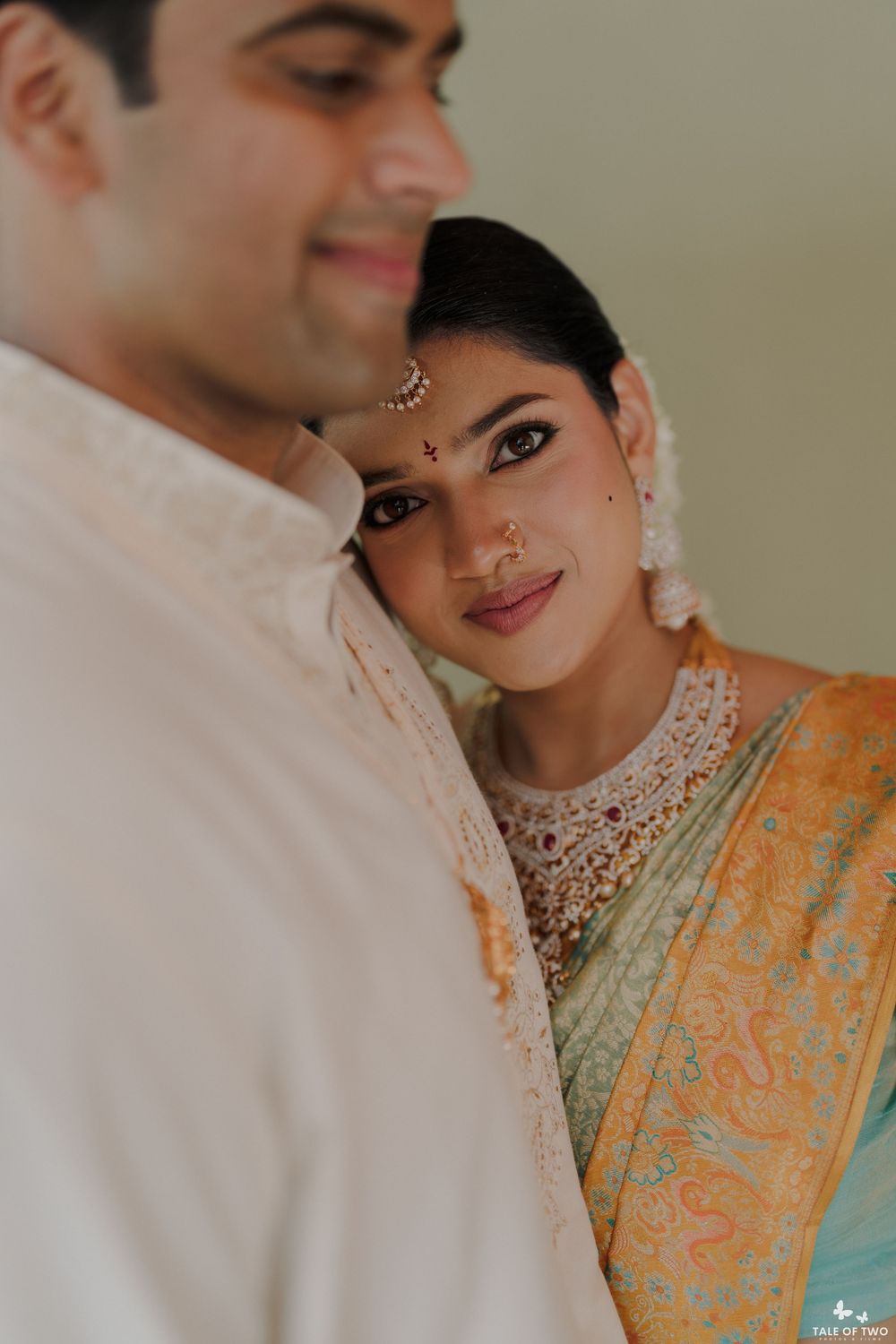 Close-up of Indian bride in teal saree with maang tikka and nose ring leaning on groom in white.