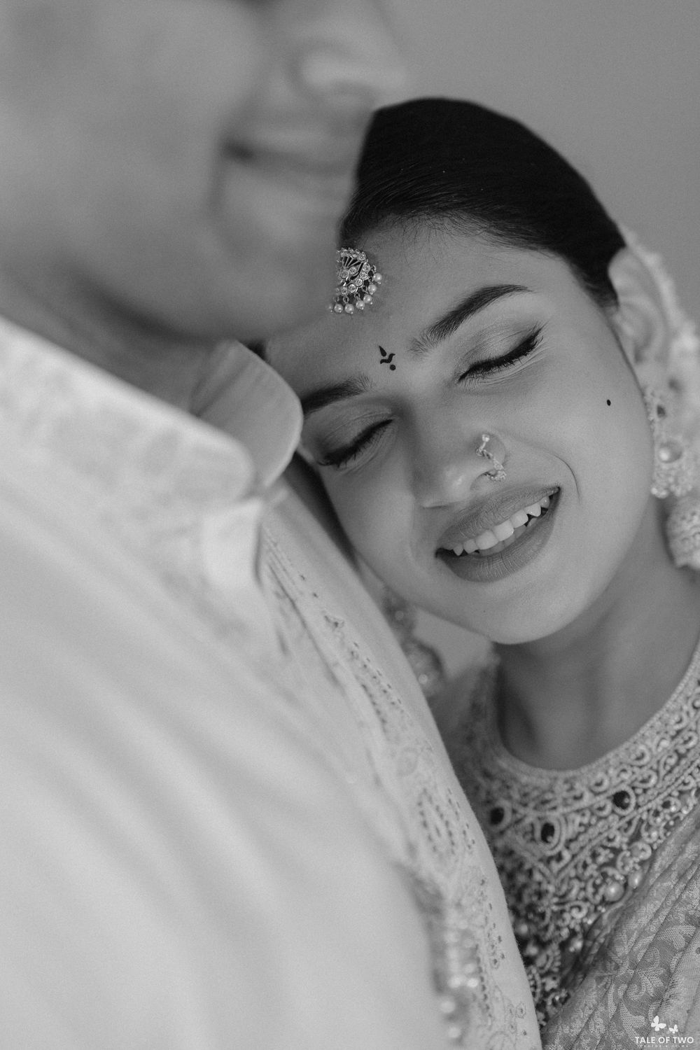 Black and white photo of a smiling Indian bride with maang tikka and nose ring leaning on groom during wedding.