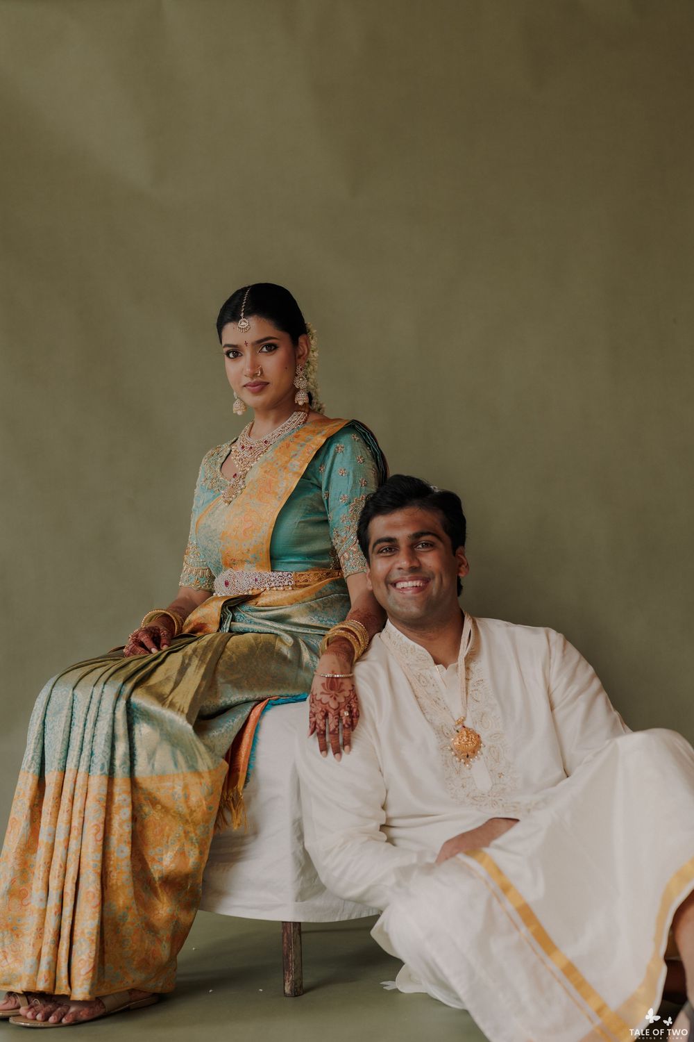 Indian bride in teal silk saree and groom in white dhoti pose elegantly for traditional wedding portrait.