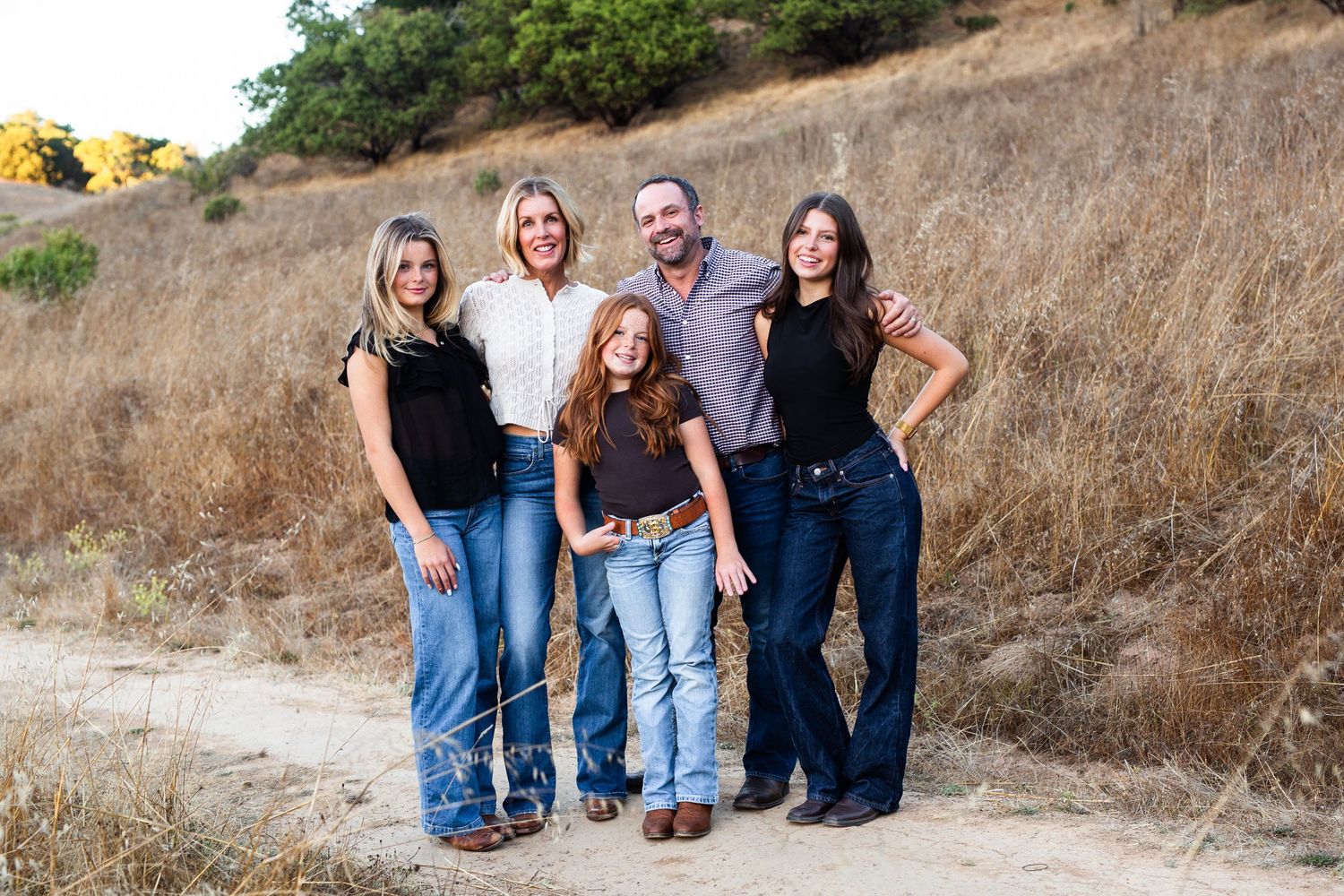 Family of five smiling outdoors on a dirt path with golden hills and green trees in the background.