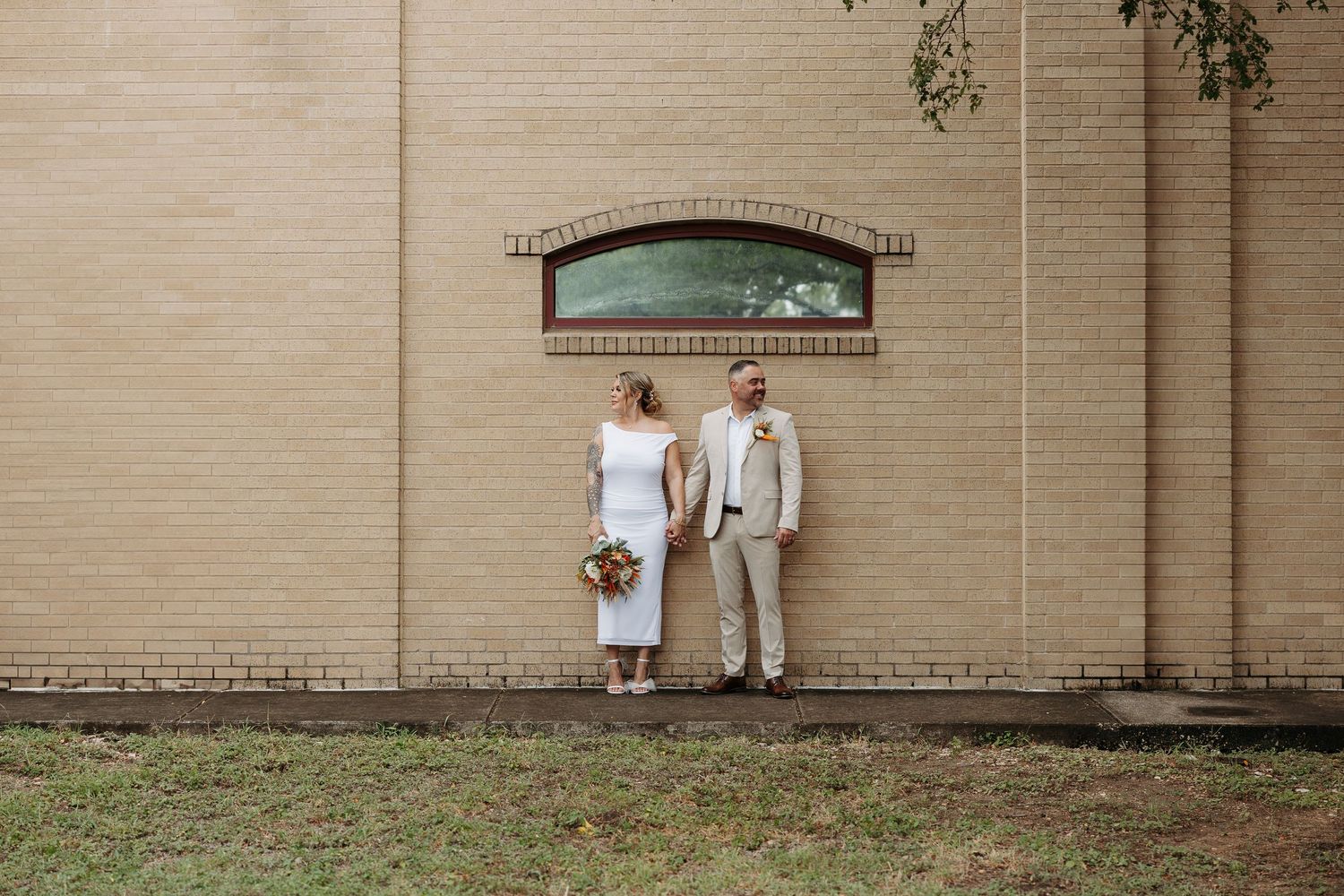Bride and groom standing apart against a tan brick wall outdoors on their wedding day.