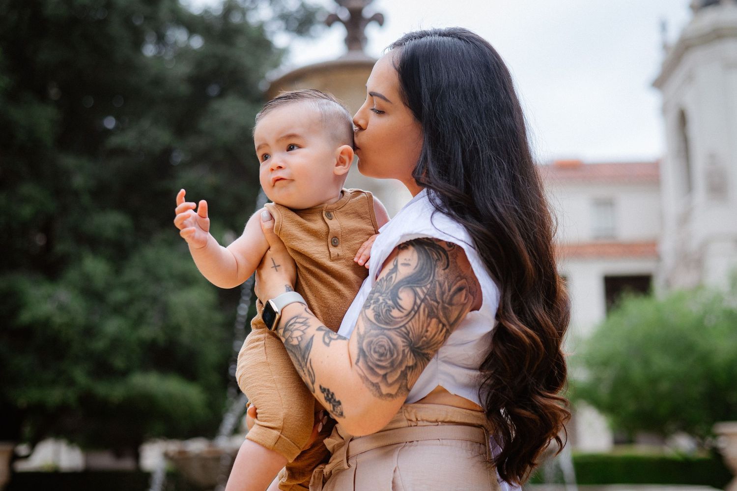 Parent holding baby in brown outfit outdoors near garden setting.