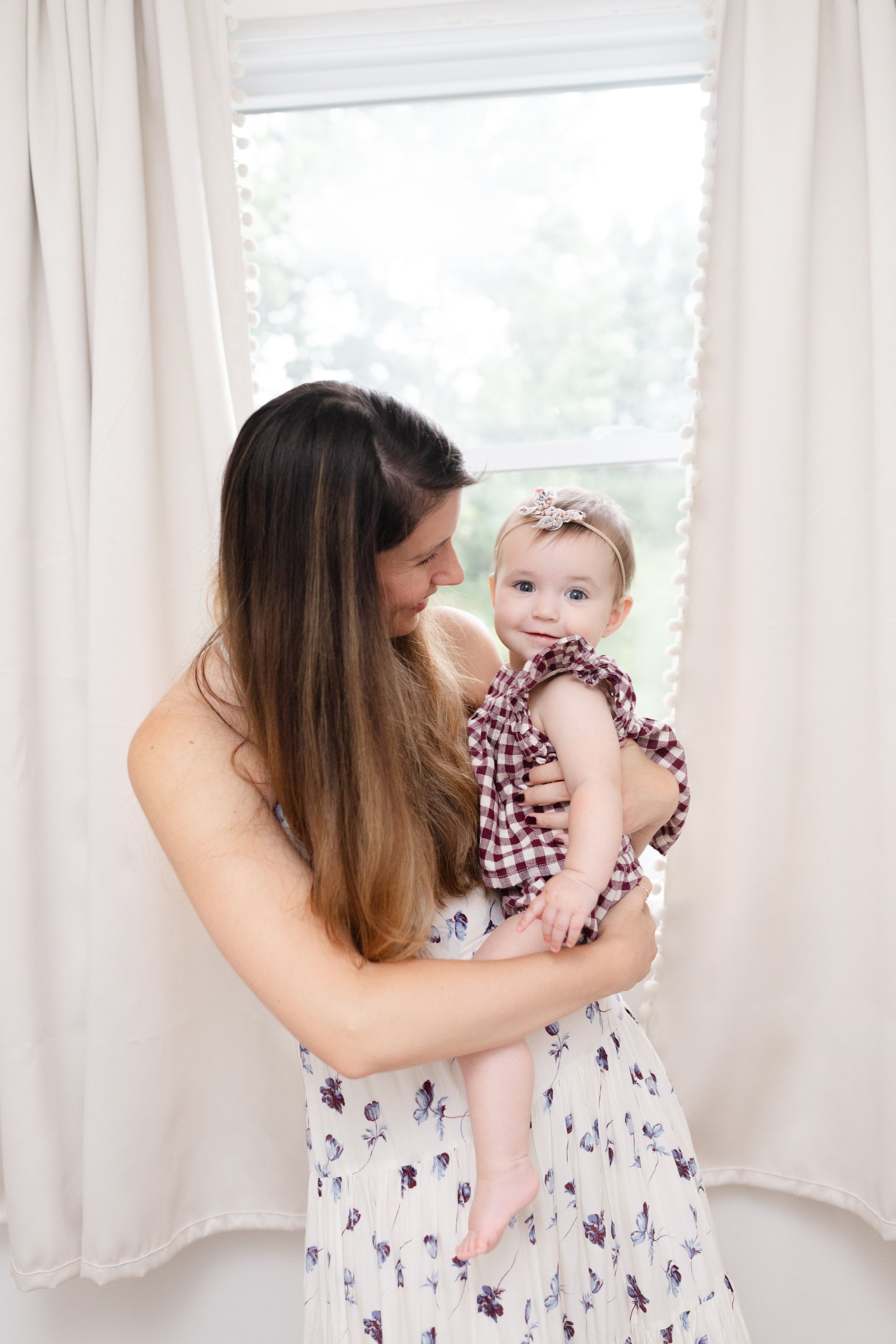A mother in a floral dress holds her baby girl in a plaid outfit near a bright window.