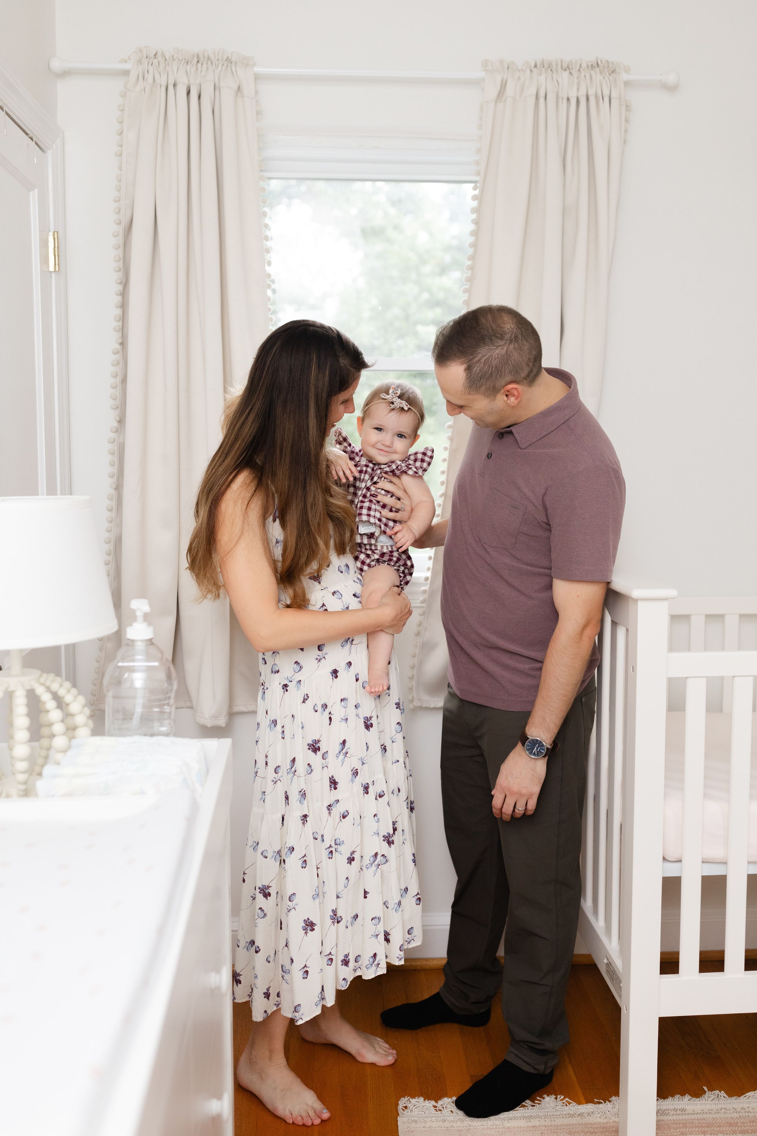 Young parents stand by white crib holding smiling baby girl in floral nursery room with natural light.