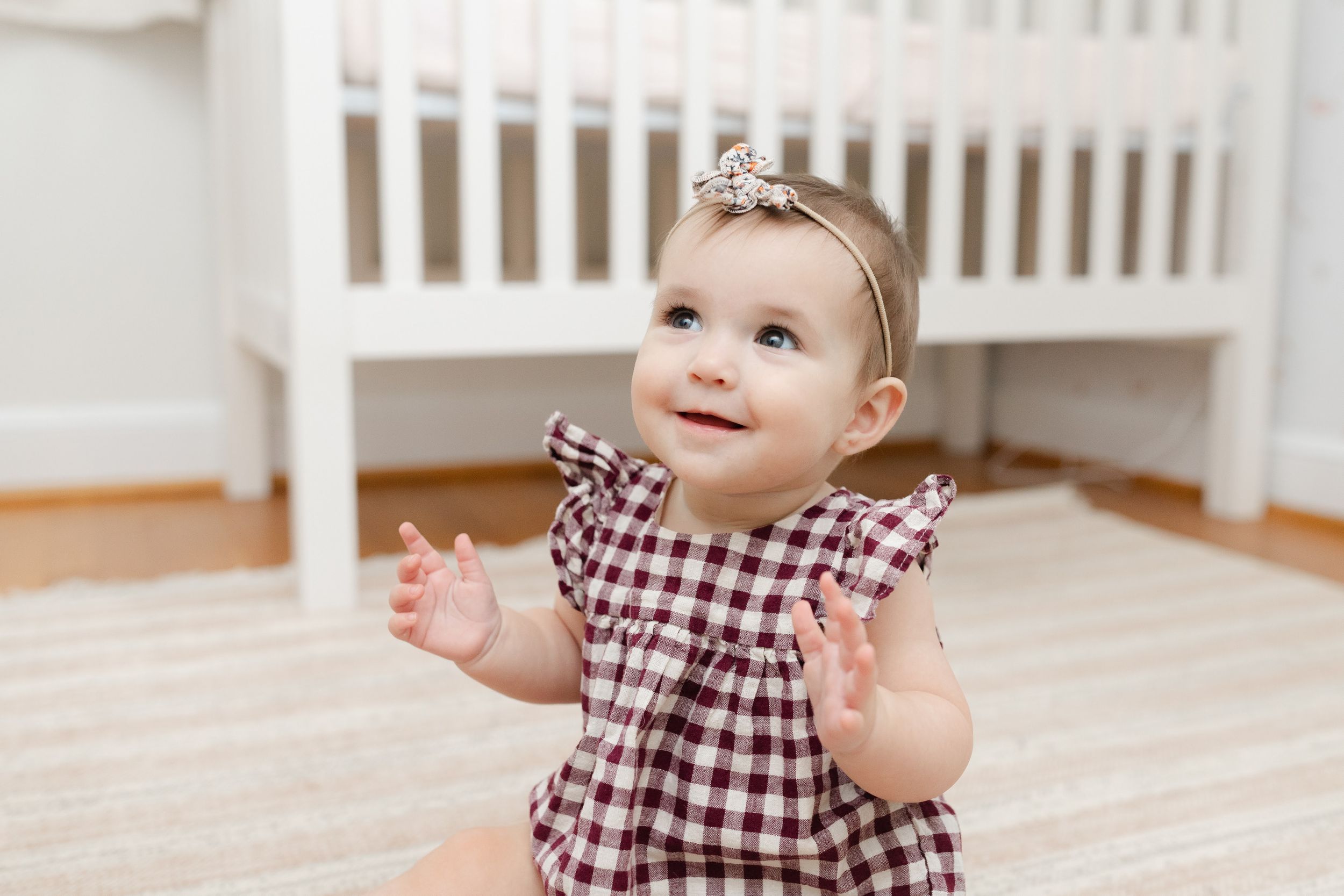 Smiling baby girl in plaid dress and floral headband clapping hands while sitting on floor near white crib.