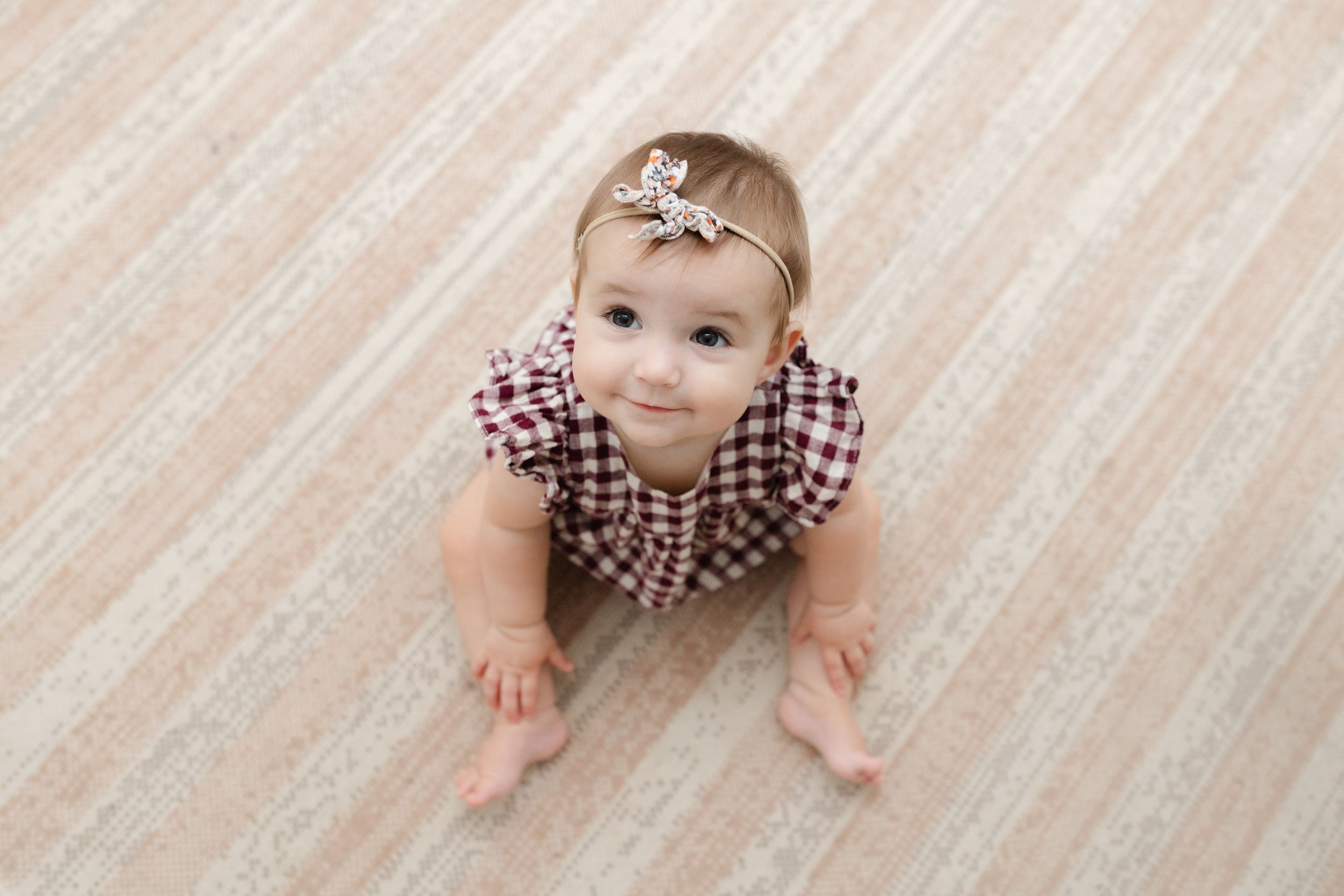 Adorable baby girl in plaid romper and bow headband sitting on light wood floor looking up at camera.