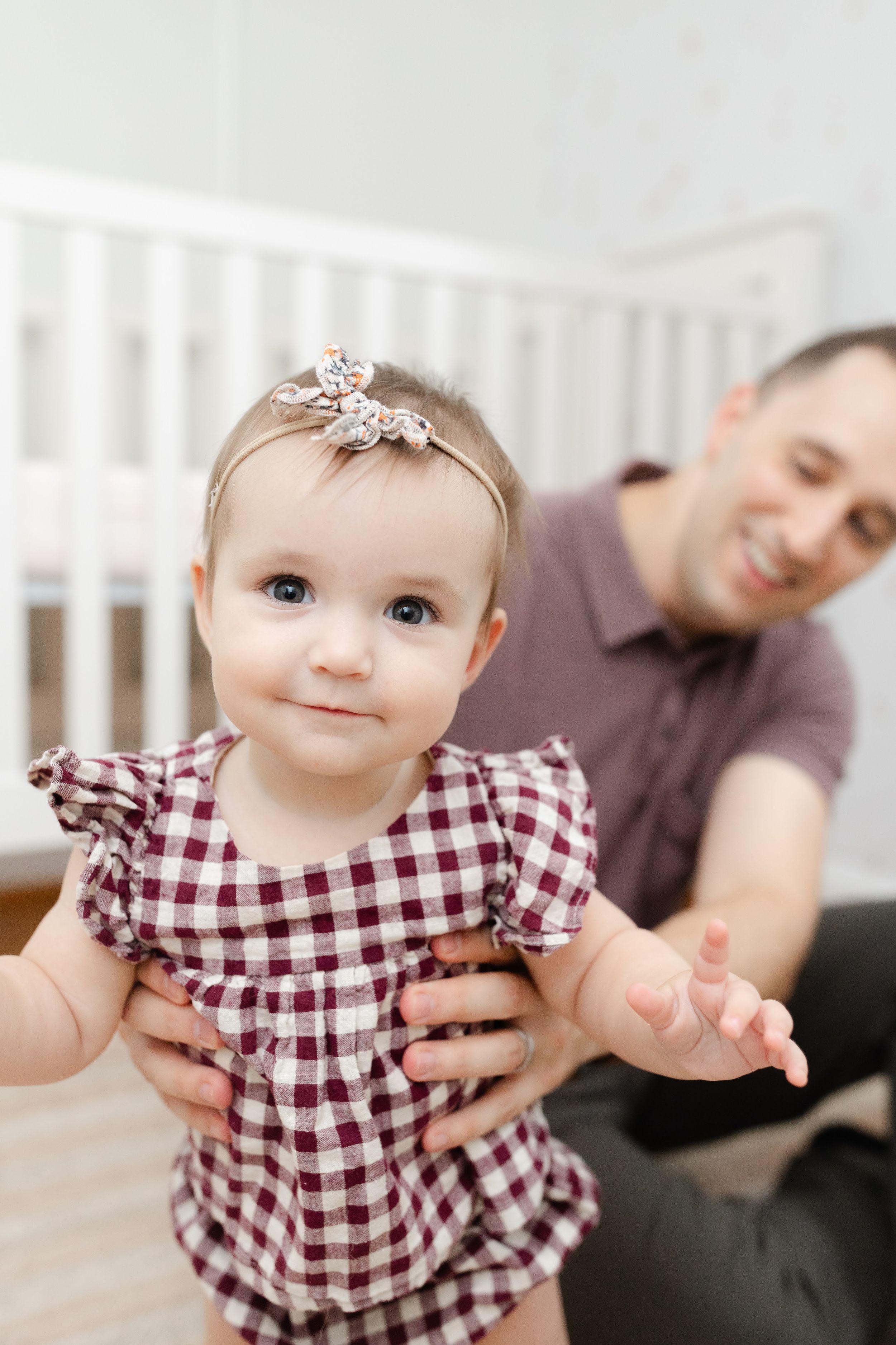 Smiling baby girl in plaid dress with floral headband takes first steps with father's support in nursery.