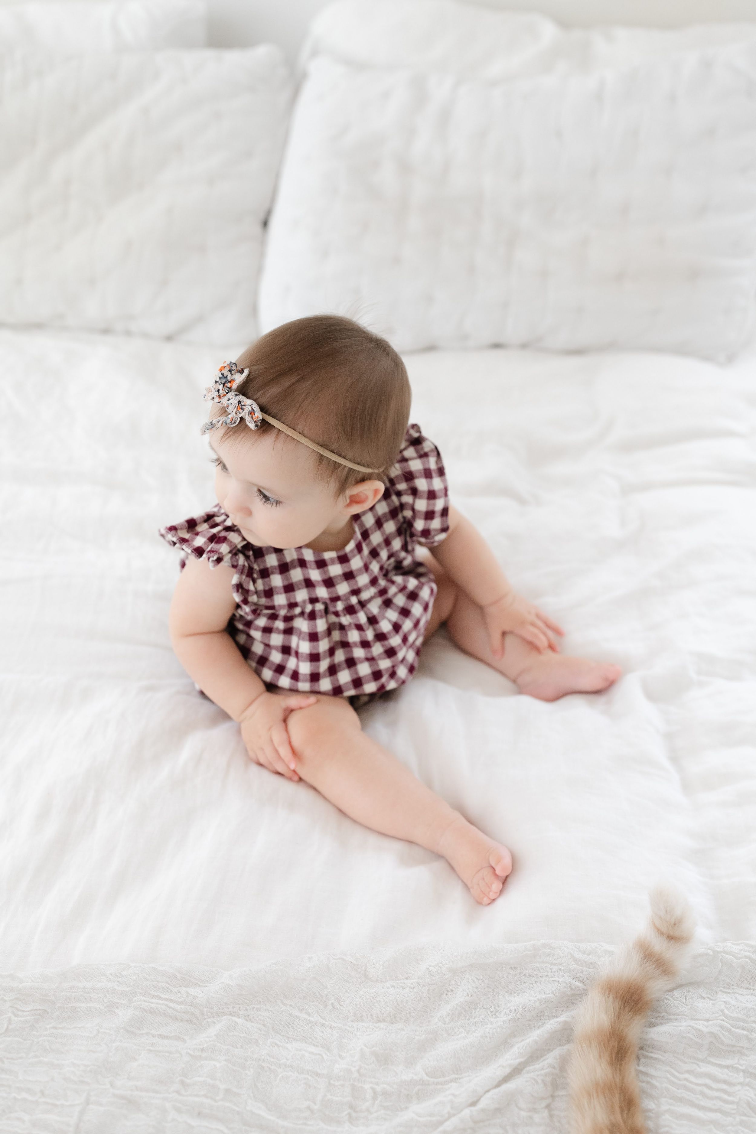 Adorable baby girl in red plaid romper and floral headband sitting on white bedding.