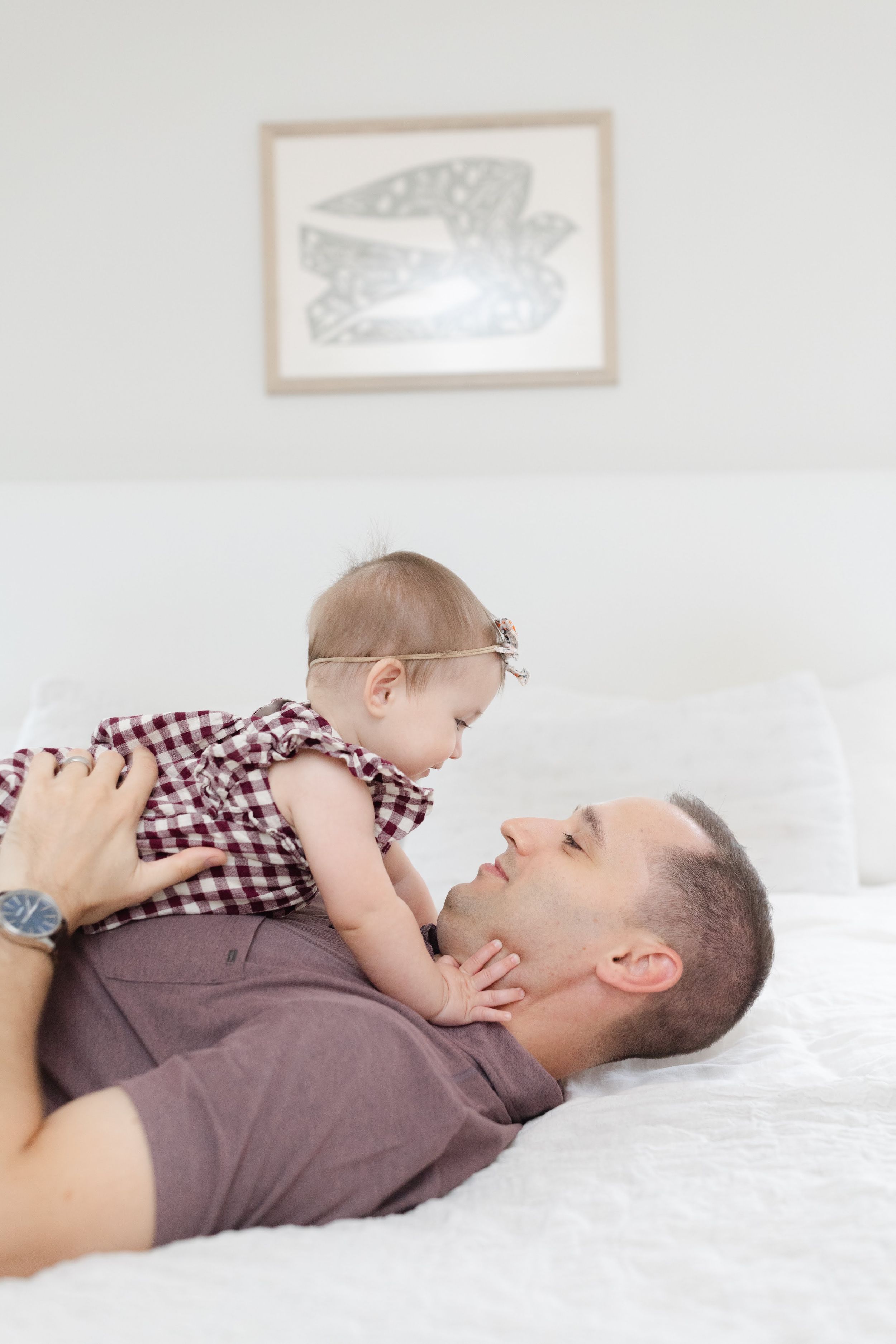 Father lying on white bed playfully holding his baby girl in red plaid dress.
