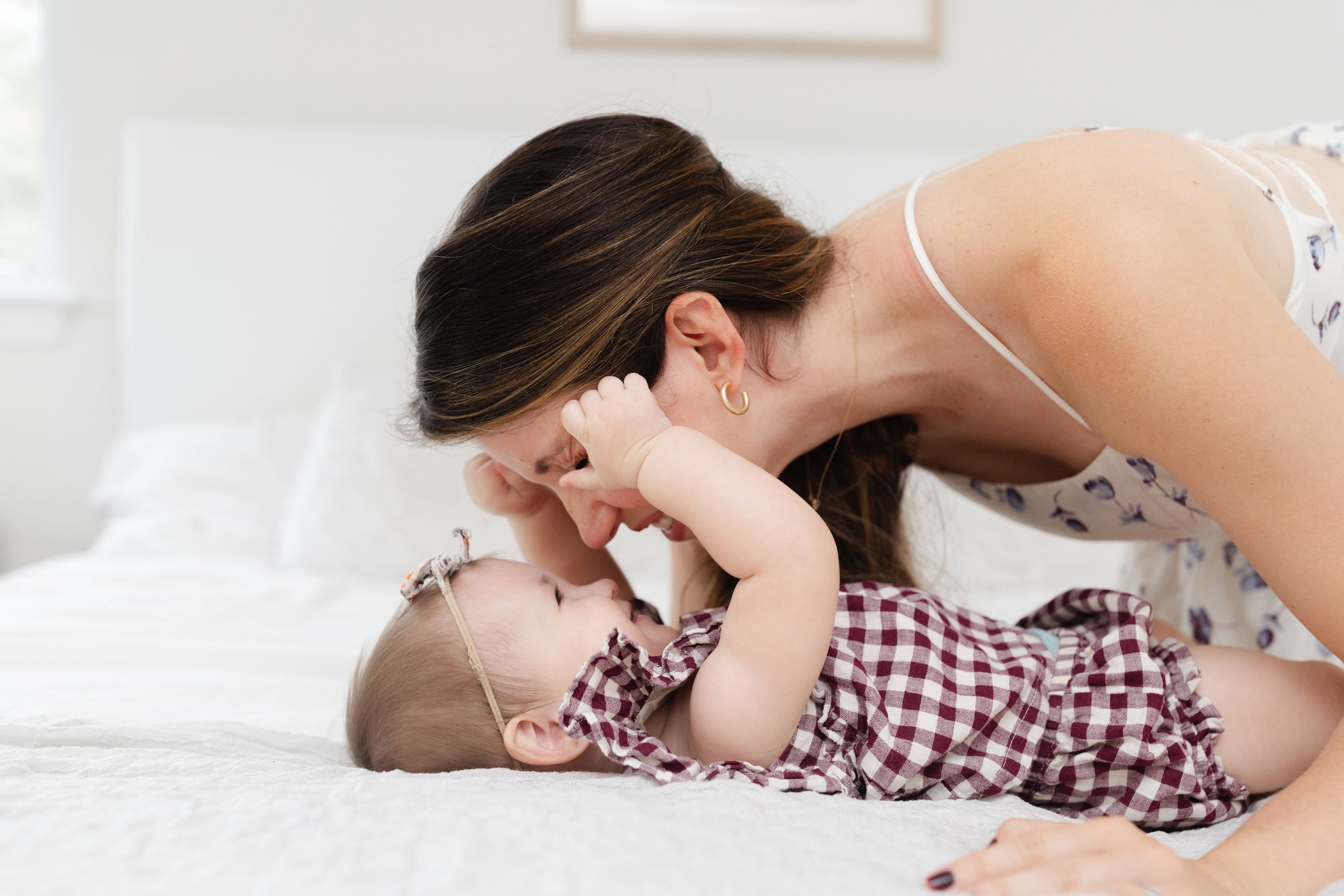 A loving mother leaning down to kiss her smiling baby lying on a white bed.