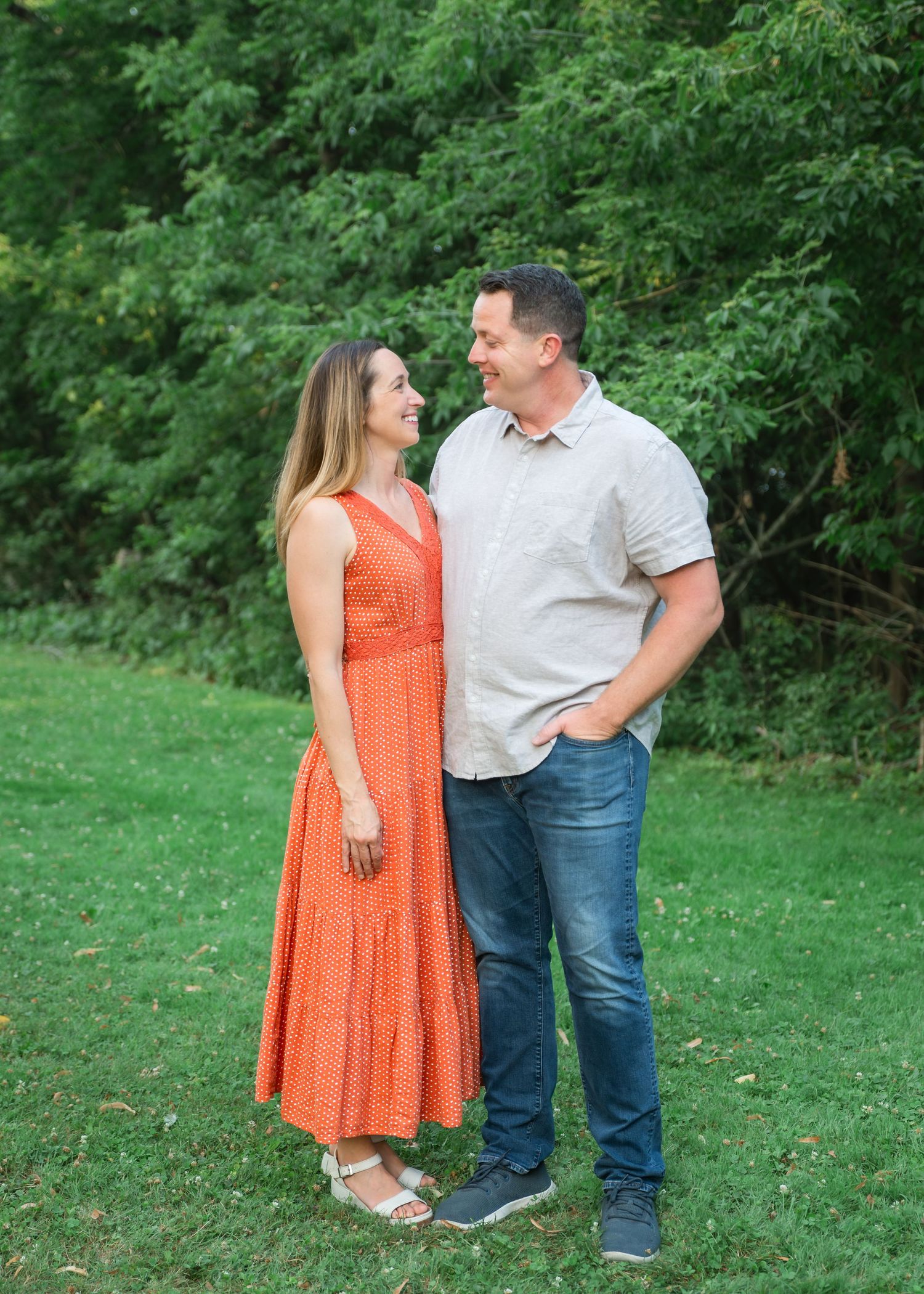 A smiling young girl in a blue dress hugs an adult's arm outdoors on green grass.