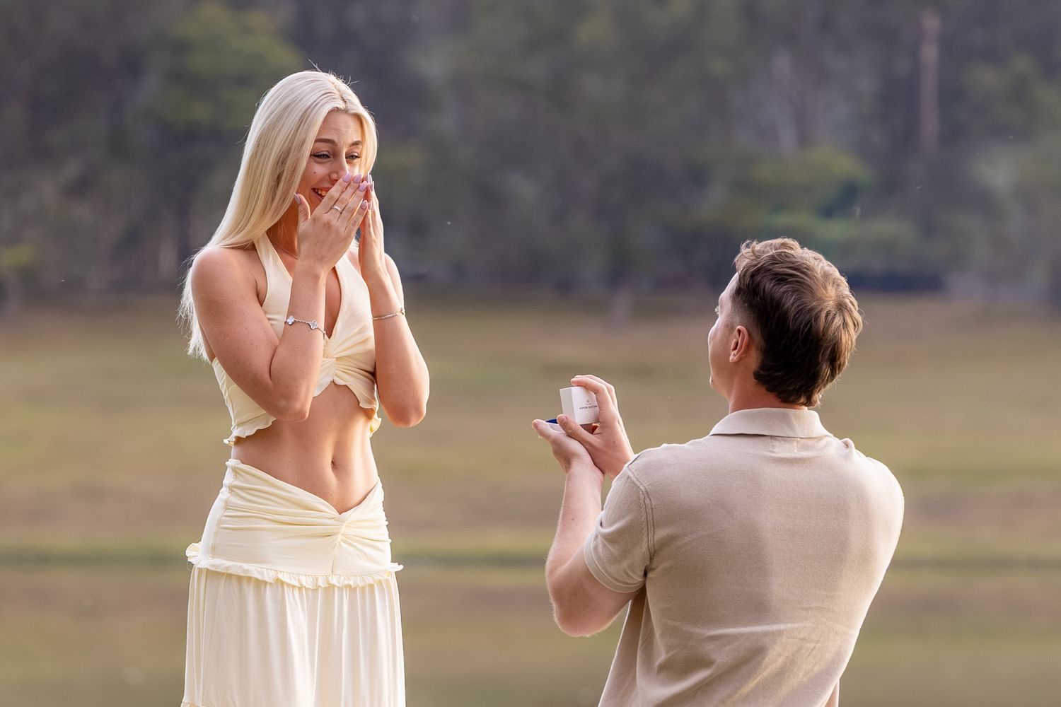 Man kneeling on one knee proposing to a surprised blonde woman outdoors near a lake with a ring box.