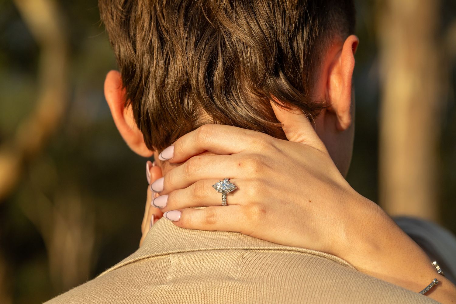 Woman's hand with diamond engagement ring resting on man's neck in golden sunlight.