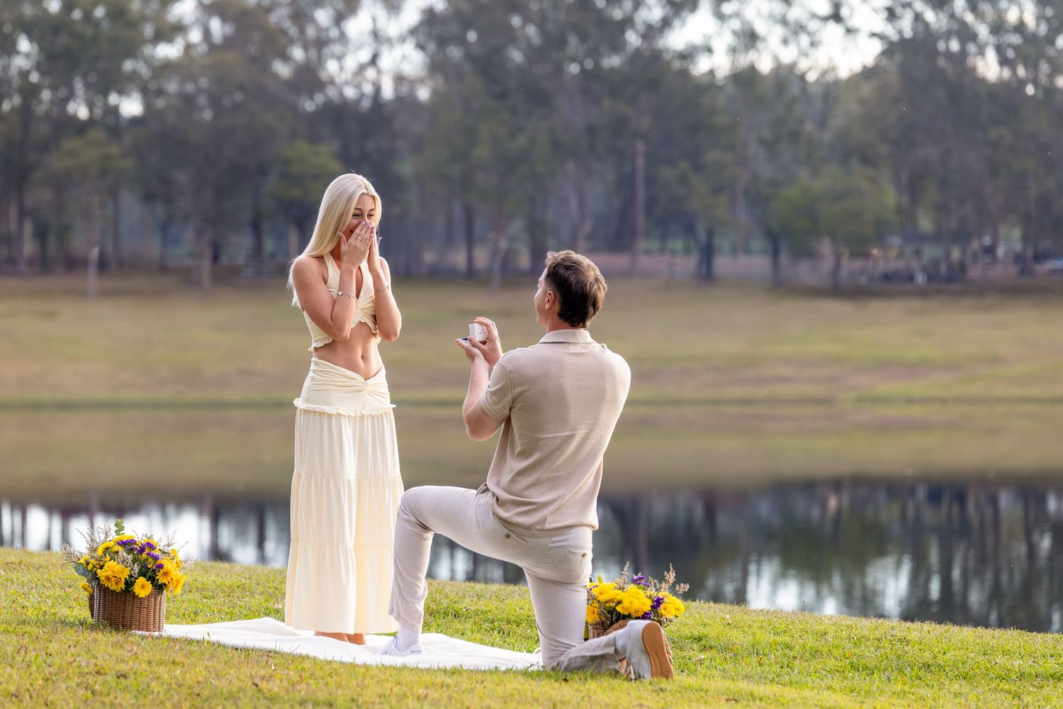 Man kneeling on grass proposing to woman near a lake with yellow flowers, romantic outdoor marriage proposal.