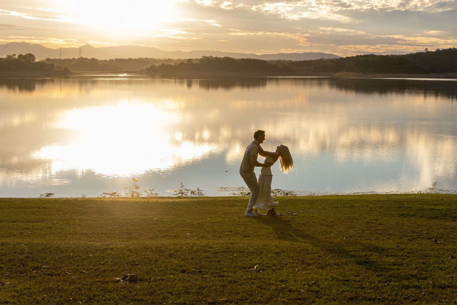 Couple dancing romantically by a serene lake at golden sunset, silhouetted against glowing water reflections.